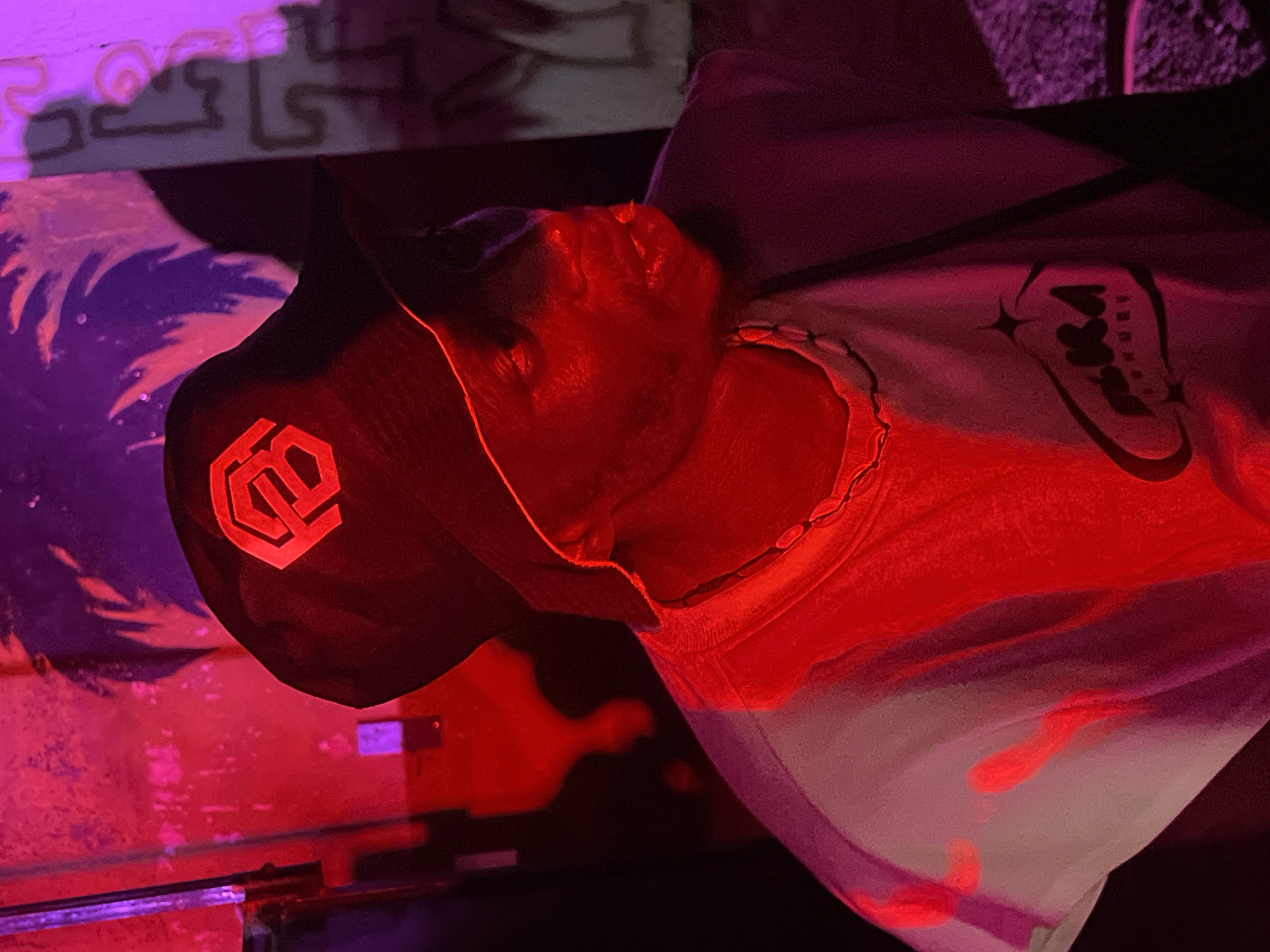 A person wearing a black bucket hat with a red emblem and a white t-shirt with a black graphic is shown in a dimly lit environment with red and purple lighting. The person's face is partially obscured and illuminated by the red light, giving it a surreal glow. The background consists of a wall with a graphic design, casting shadows. The overall impression is one of a stylized portrait taken in an urban setting, possibly a club or art space. The location in Durimi, Nigeria, is not visually identifiable in the provided image.