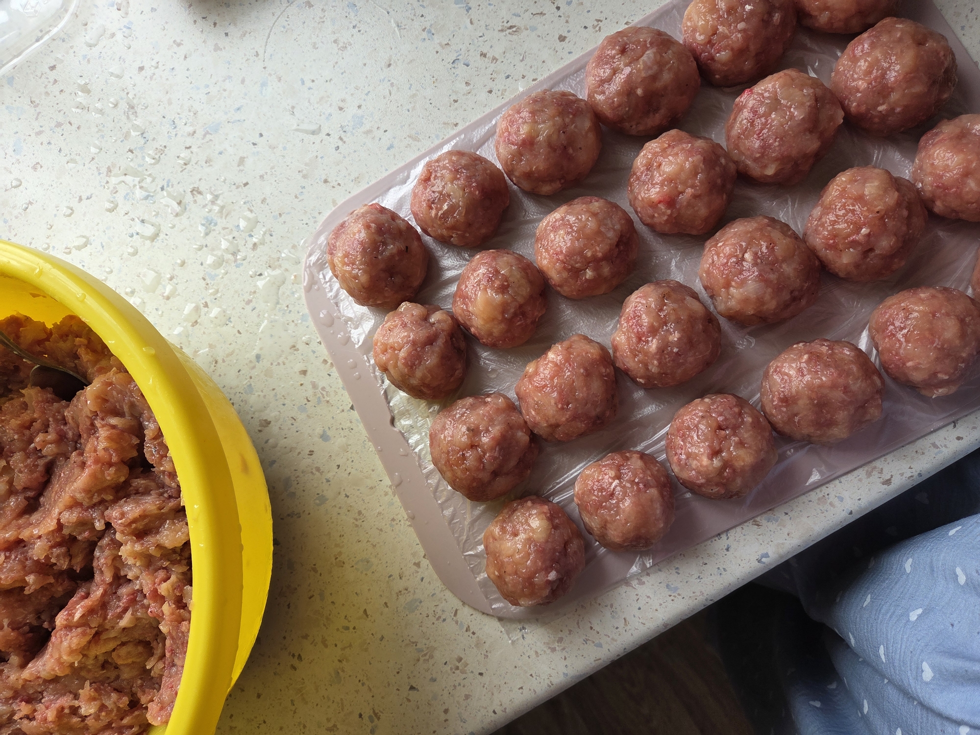 A high-angle shot reveals a preparation scene involving raw meatballs. A yellow bowl, partially filled with ground meat mixture, occupies the left side of the frame. To the right, on a pink cutting board lined with plastic wrap, are approximately 25 neatly arranged, uncooked meatballs. Droplets of water are scattered on the countertop. A portion of a light blue garment with small white heart pattern is visible in the lower right corner. The image appears to be taken indoors, possibly in a kitchen. There is no information available to suggest this scene is taking place in Birobidzhan, Russia.