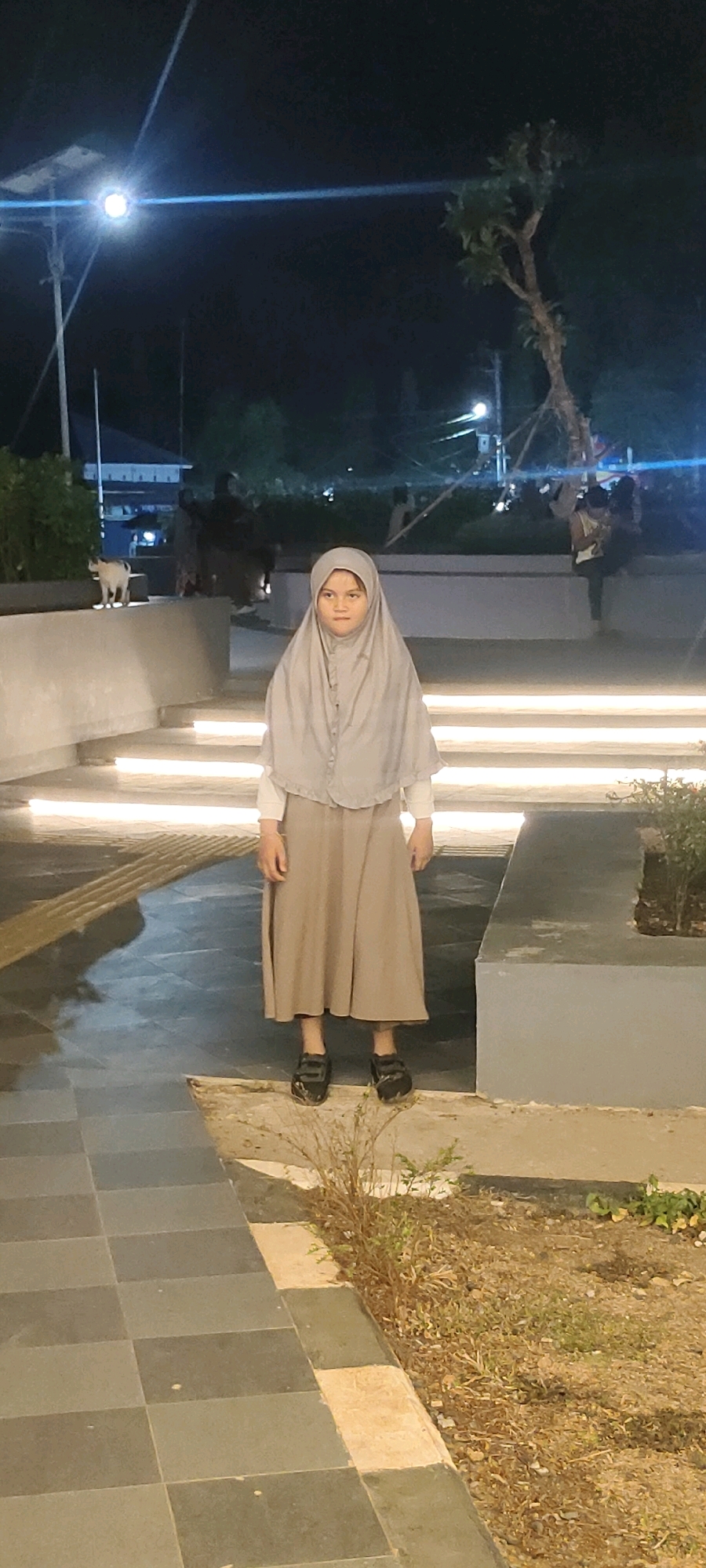 A young girl wearing a light grey hijab and a long, flowing, light brown skirt stands on a paved area at night. She is looking directly at the camera with a neutral expression. The area appears to be an outdoor public space, possibly a park or plaza, with modern lighting integrated into the steps and pathways. In the background, trees, streetlights, and silhouetted figures are visible. The lighting suggests it is after sunset. The girl is standing on a slightly elevated, concrete platform adjacent to a tiled walkway. The environment suggests it is a well-maintained urban outdoor space.