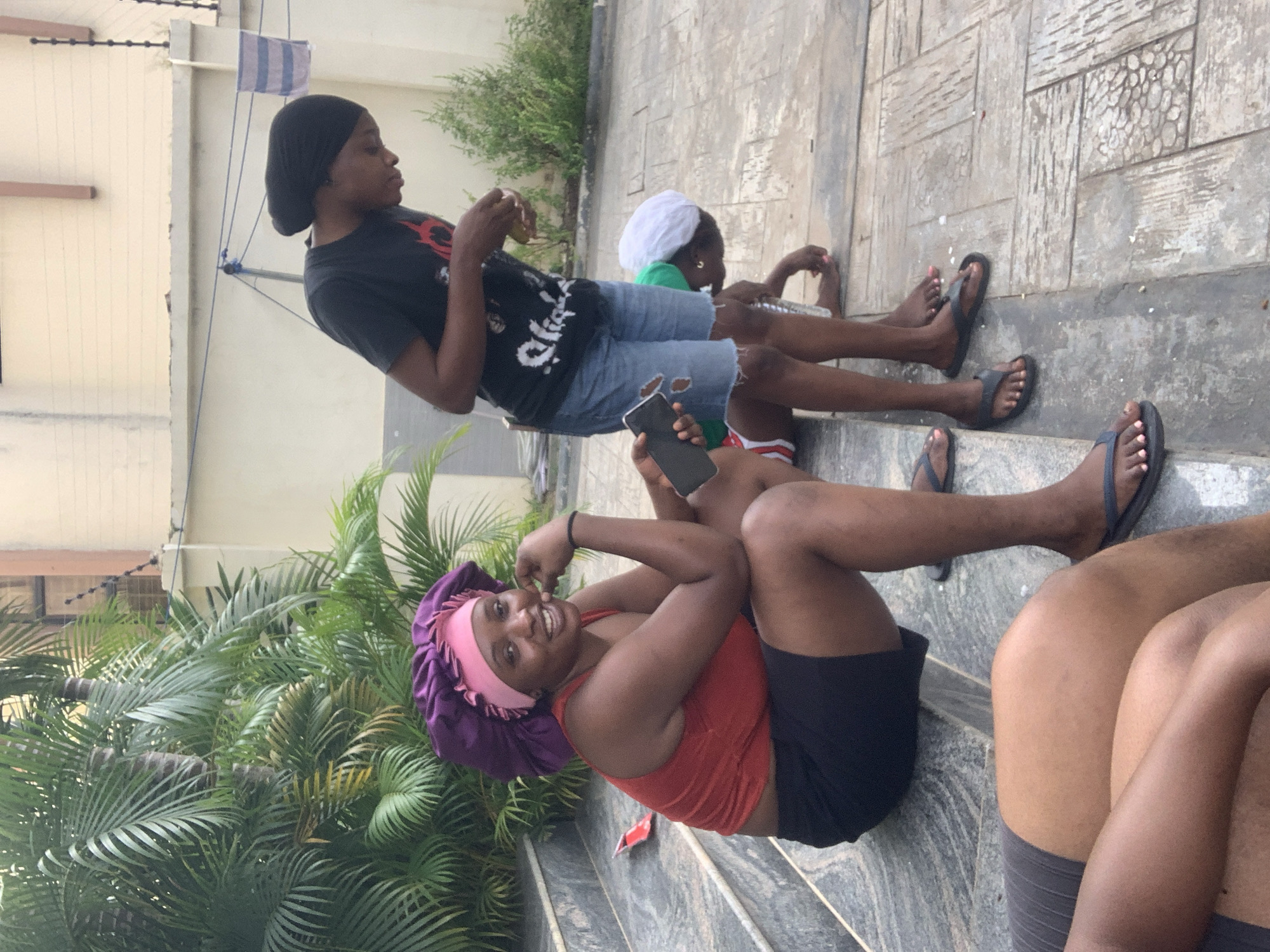A group of individuals are casually gathered outdoors on a paved patio with some greenery in the background. The scene appears to be in Abuja, Nigeria.
In the foreground, three individuals are visible. On the left, the legs and lower torso of one person are shown, wearing dark shorts. Next to them, a woman is seated on stone steps, wearing a red tank top and black shorts, with a purple and pink bonnet on her head. She is smiling and holding a black smartphone in her right hand. To her right, another person, possibly a young individual, is seated on the ground, wearing a green shirt and a white cap. Further back, a taller individual stands, wearing a black t-shirt and denim shorts, and appears to be holding something in their hands. They are looking downwards.
The environment includes stone steps, a tiled patio, lush green palm trees, and a pale-colored building in the background with a black security fence on top. A clothesline with a striped towel is also visible on the building. The overall atmosphere suggests a relaxed, informal gathering.
