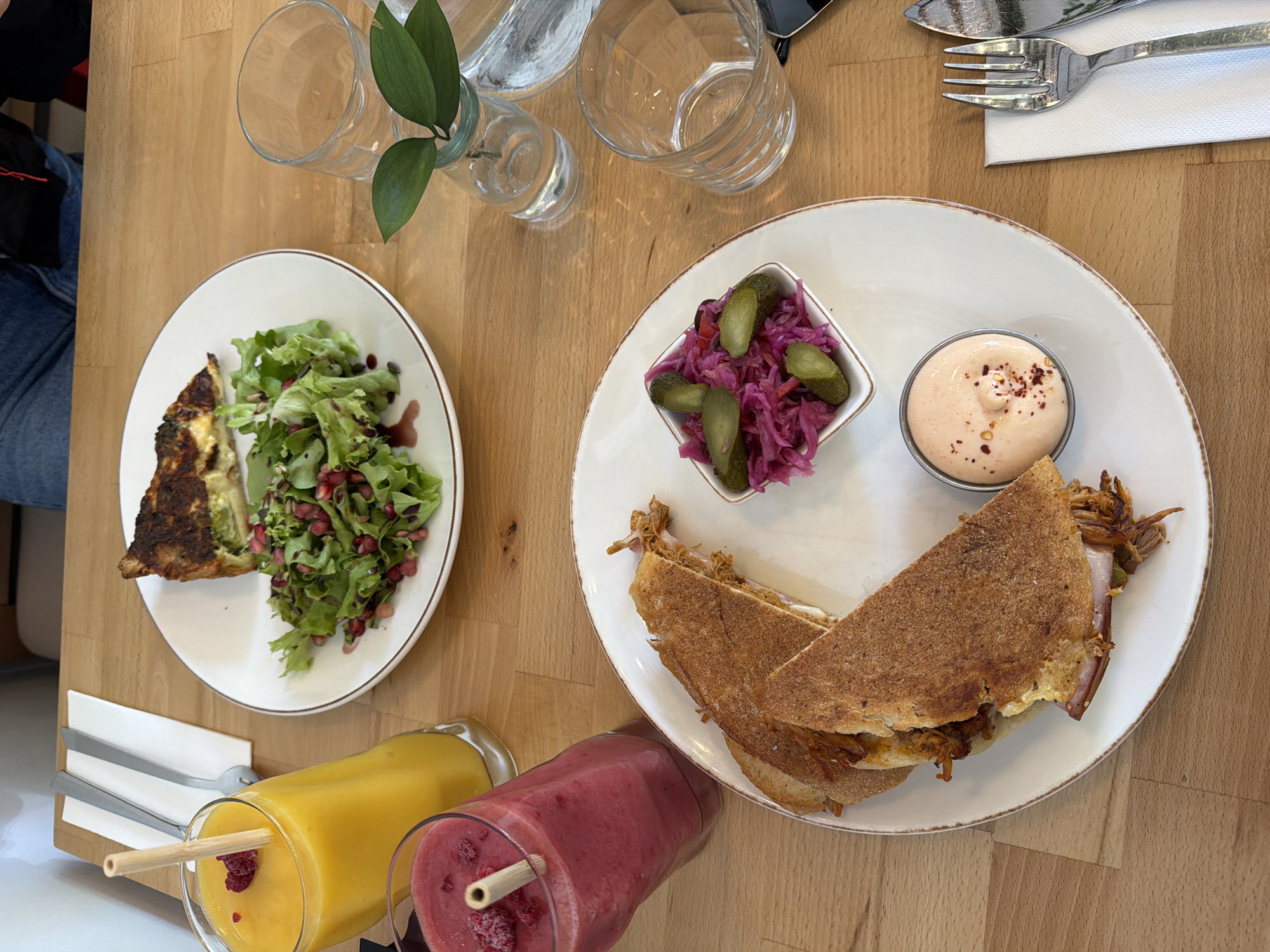 An overhead shot depicts a wooden table in a restaurant setting, likely in Sofia, Bulgaria, evidenced by the style of cuisine often found in Sofia's cafes and restaurants. The table features two white plates, each holding a distinct meal. The plate on the left contains a slice of quiche accompanied by a green salad speckled with pomegranate seeds. The plate on the right presents a toasted sandwich paired with a small dish of pickled cabbage and gherkins, and a ramekin of pinkish sauce topped with red pepper flakes. Two beverages, one orange and one red, in tall glasses with straws are positioned near the bottom edge of the table. Cutlery, wrapped in a white napkin, rests near the top edge of the table. A small vase with green foliage sits centrally between the two plates. Two empty drinking glasses and a water bottle are arranged behind the vase. A partial view of a person wearing blue jeans is visible on the left edge of the image. The scene suggests a casual dining experience in a well-lit indoor environment.