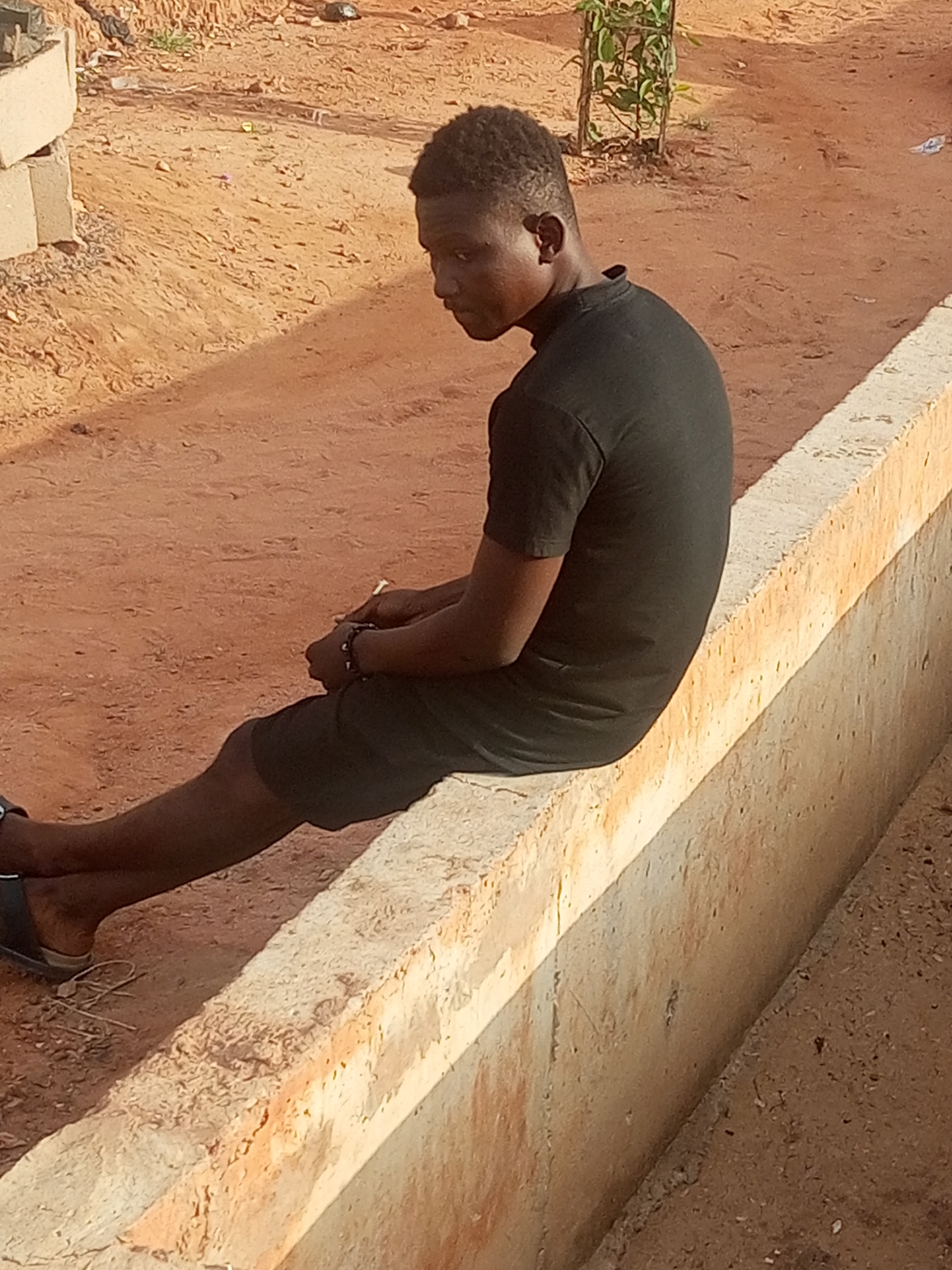 In a dusty, outdoor environment, a young Black male is seated on a raised concrete ledge, facing to the right and looking over his left shoulder. He is wearing a black t-shirt and dark shorts, with dark sandals on his feet. His hands are clasped in front of him, and he has a bracelet on his left wrist. The background consists of reddish-brown dirt with some scattered rocks and a small green plant in a planter behind him. A pile of concrete blocks is visible to the far left. The ledge he is sitting on is weathered and appears to be part of an outdoor structure, possibly a planter box or a low wall. The lighting suggests it is daytime, with shadows cast on the ground.
