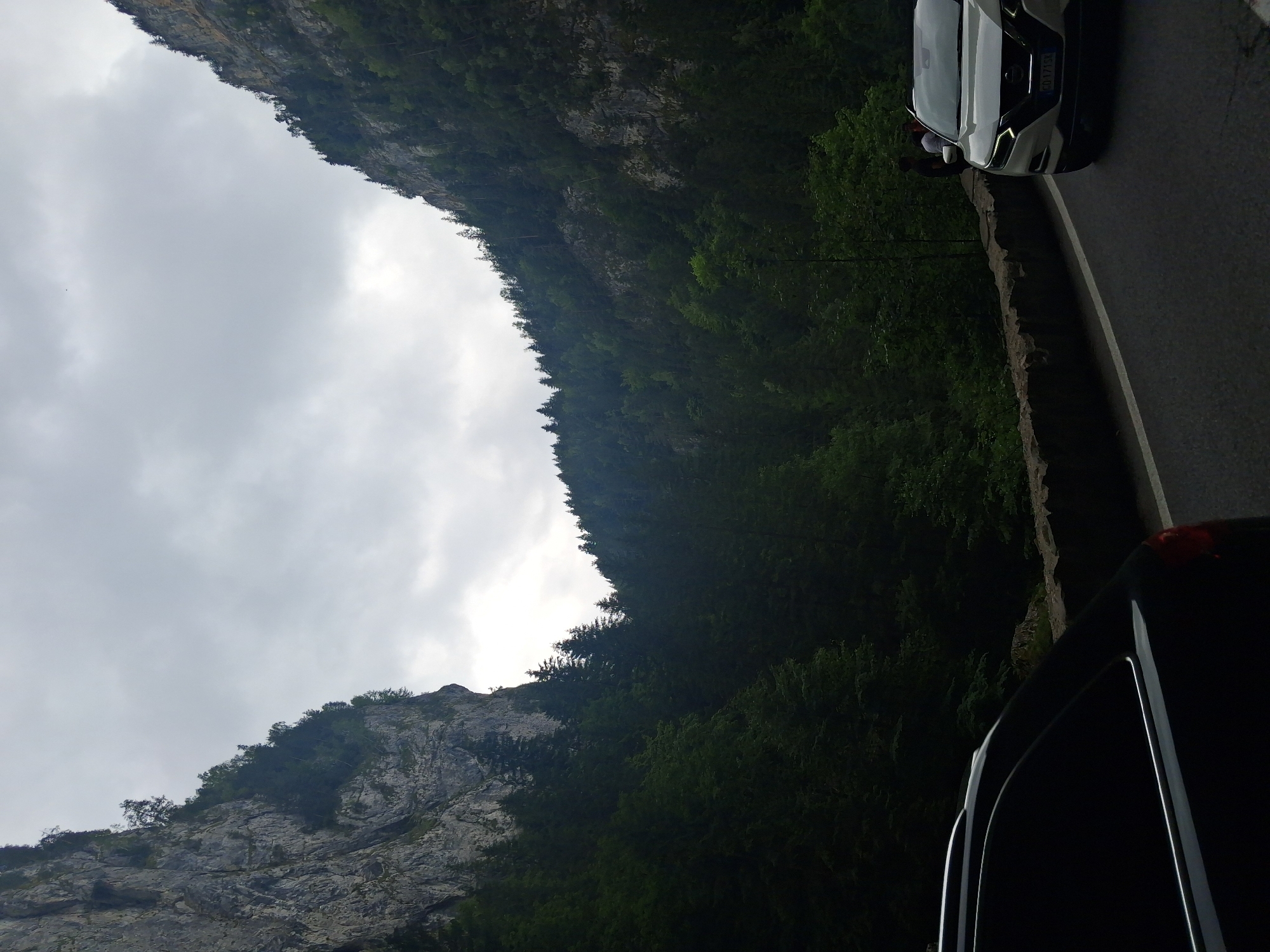 The image shows a view of a mountain road in Bicaz-Chei, Romania, with steep, rocky cliffs covered in dense forest on either side. The sky is overcast, with dark clouds creating a dramatic atmosphere. In the foreground, the rear of a black car is visible on the left, and on the right, a white SUV is driving on the road. Two individuals are standing on the side of the road near the white SUV. The road is paved and has a white line marking its edge. The surrounding environment is characterized by lush green vegetation and imposing rock formations.