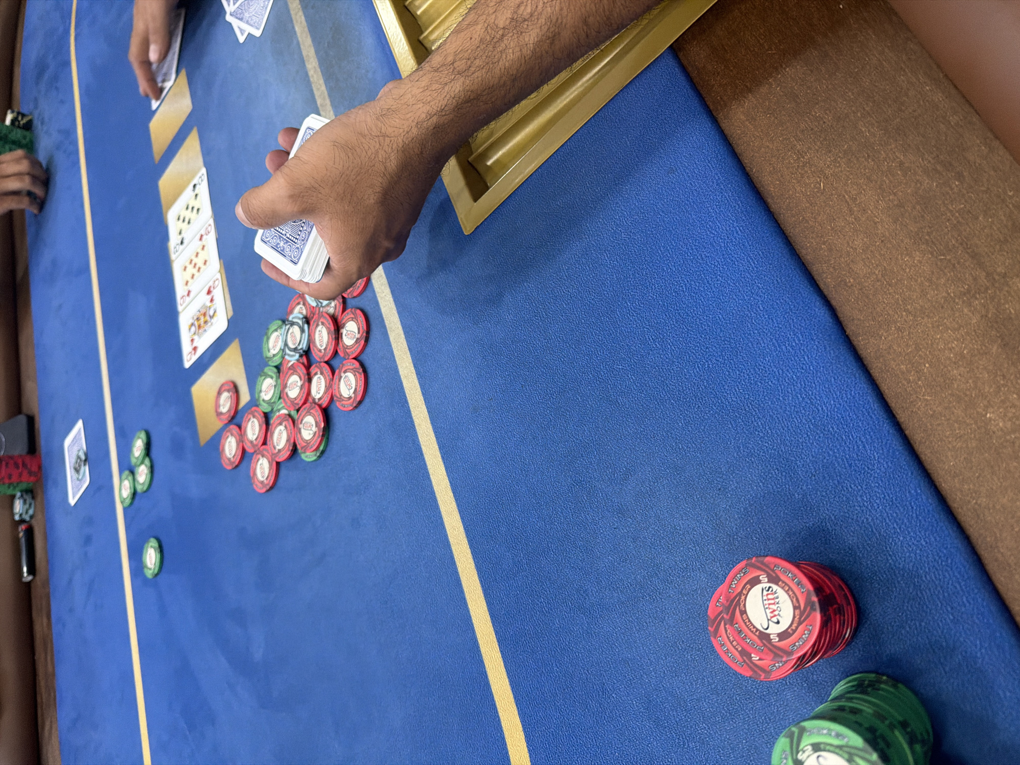 The image depicts a poker game in progress, possibly in Larnaca, Cyprus. A blue felt-covered table with a yellow dividing line dominates the scene. A player's hand is visible, dealing cards. The community cards on the table are 10 of clubs, 8 of clubs, 7 of hearts, 5 of hearts, and queen of hearts. Stacks of red and green poker chips are scattered on the table, along with a card holder.