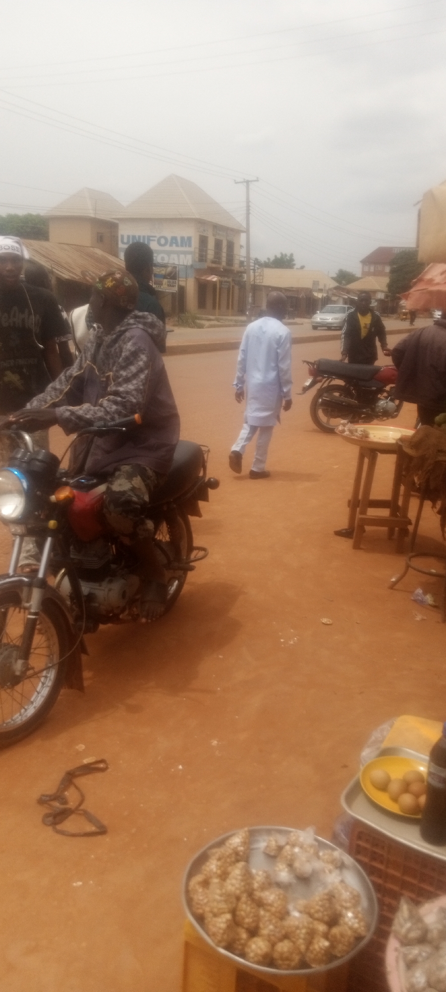 The image depicts a street scene in Sabon Tasha, Nigeria. A man on a motorcycle faces left. Another man in a white shirt and pants walks away from the camera in the center of the frame. Two motorcycles are parked in the background. Near the bottom of the frame, a tray of spherical snacks and a plate of what looks like fried eggs rest on a makeshift table, likely for sale. The ground is reddish-brown. A building with "UNIFOAM FOAM" on the facade is visible in the background. Overhead power lines cross the sky. The scene appears to be an outdoor market or trading area.