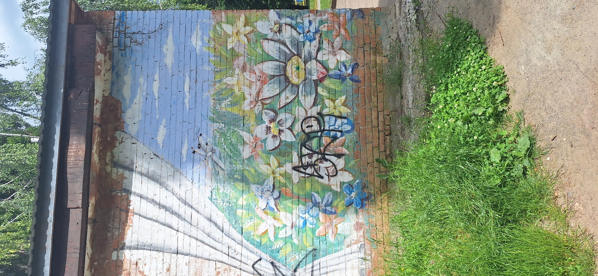 A mural painted on a brick wall depicts a sky with clouds and a field of flowers. The mural is partially obscured by overgrown green grass and a dirt path in the foreground. The top of the wall is covered by a corrugated metal roof. The mural has graffiti in black spray paint over the flowers. The scene appears to be outdoors in an urban or suburban setting, possibly in Bila Tserkva, Ukraine, given the trees and vegetation.