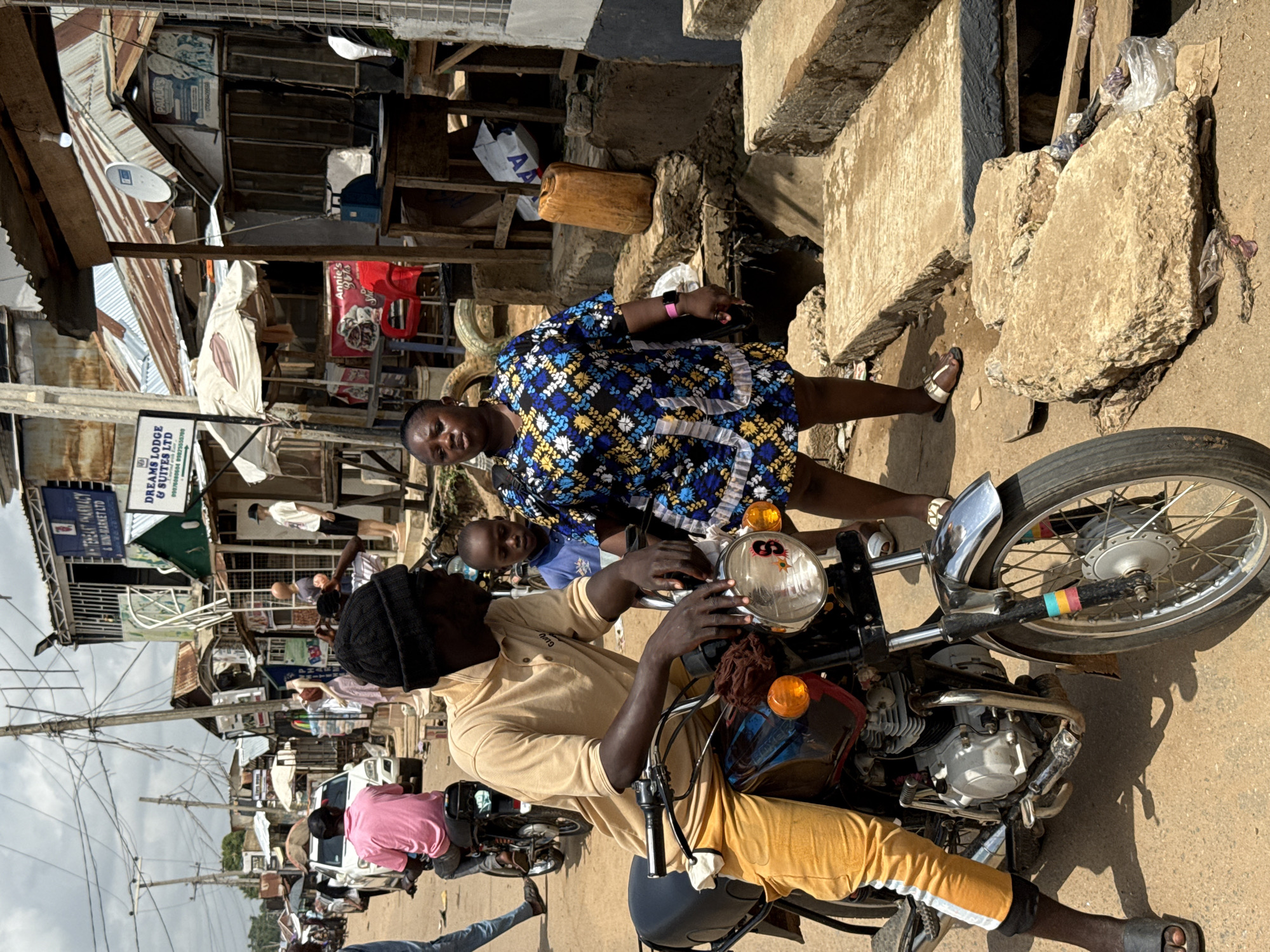 The image depicts a street scene in Abuja, Nigeria, with a motorcycle at the forefront. A man in a tan shirt and yellow pants sits on the motorcycle, while a woman in a blue and yellow dress stands next to him. A young boy in a blue shirt is positioned between them. The surrounding environment includes buildings with corrugated metal roofs, various signs (including "Dreams Lodge & Suites Ltd"), and a partially visible concrete structure. People are seen walking in the background, along with other motorcycles. Utility poles with overhead wires are visible against the sky. The scene suggests a bustling, informal urban environment.