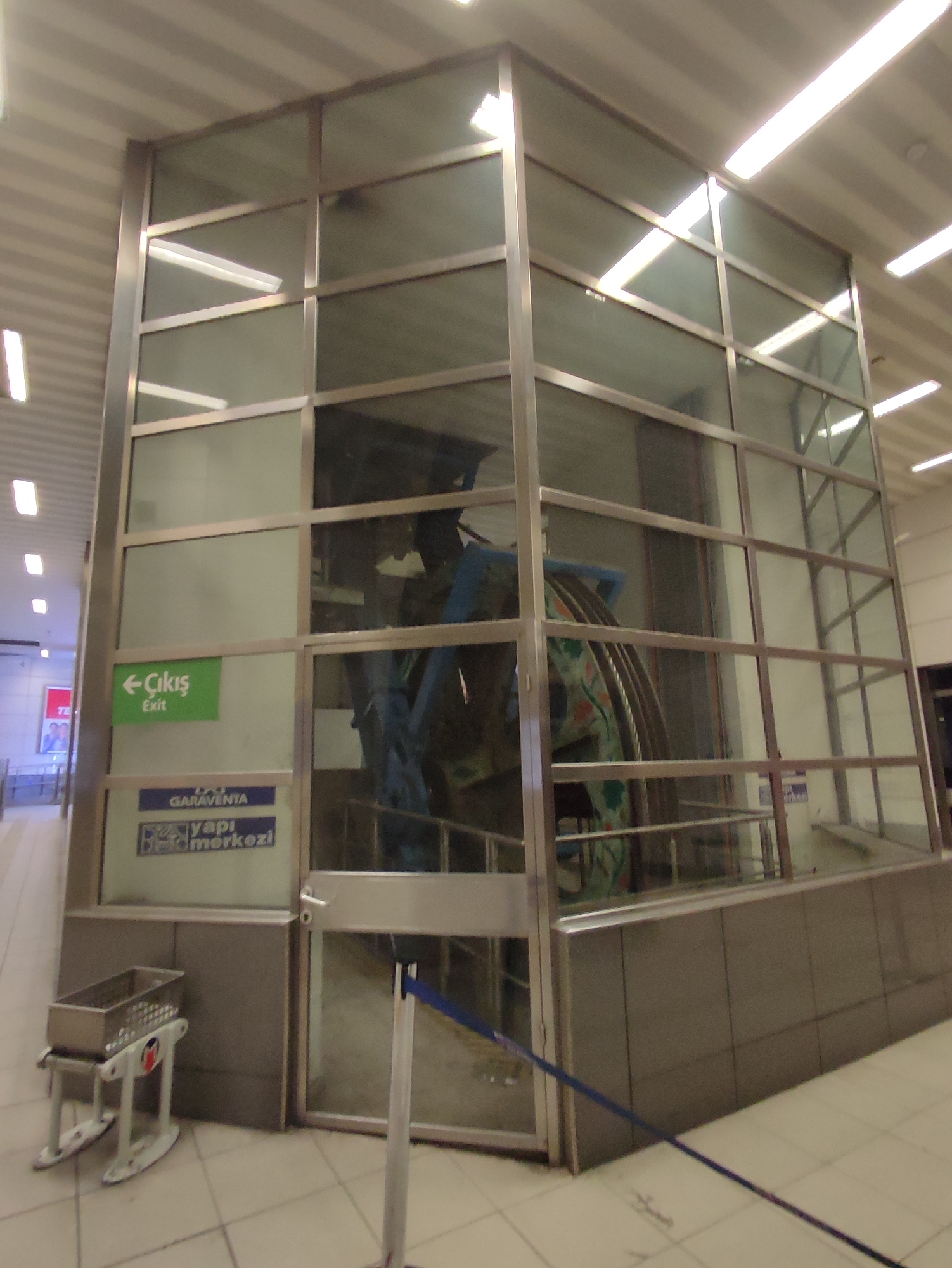 The image shows a modern glass-enclosed structure, likely an elevator or funicular, in an indoor, possibly public, space. The structure has a metal frame with large glass panels, allowing a view of the machinery and cables inside. A green sign with Turkish text, reading "Çıkış" (Exit) and "Exit" in English, is visible on the left side of the glass enclosure. Below that, a blue and white sign reads "GARAVENTA" and "yapı merkezi". In the foreground, to the left, is a small, wheeled metal basket, possibly for luggage or shopping. A blue rope barrier extends across the lower right portion of the image. The overall environment appears to be clean and well-lit, with fluorescent lighting fixtures visible on the ceiling. The style of the structure and the signage suggest a modern transportation hub or public facility. The location is identified as Beyoğlu, Turkey.