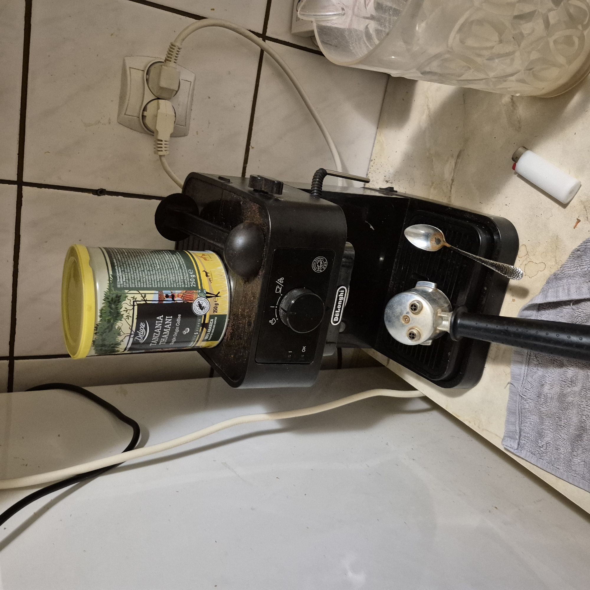 The image depicts a close-up, eye-level shot of a domestic kitchen counter in Kępno, Poland. The counter is a light-colored laminate with visible stains and dirt, suggesting it is a used surface. Against the wall, which is tiled with white, square tiles, a black De'Longhi espresso machine is the central focus. The machine appears somewhat dusty. On top of the machine rests a cylindrical can of "Deluxe Tanzania THAMANI Single Origin Coffee," also appearing dusty. To the right of the coffee can, a black lever or handle is visible, likely part of the espresso machine's design.
In front of the espresso machine's main body is the portafilter, a metal component with a black handle, positioned as if it has just been used or is about to be used. Next to the portafilter, a tarnished silver spoon lies on the counter. To the right of the espresso machine, a clear plastic pitcher or carafe is partially visible, filled with a light-colored substance, possibly water or ice. Further to the right, on the counter surface, a white disposable lighter is present.
On the left side of the image, a white vertical appliance or wall is visible, with a black electrical cord and a white electrical cord running down it. Another white electrical cord connects to a white double-plug socket mounted on the tiled wall behind the espresso machine. A grey towel is folded and placed on the edge of the counter in the lower left portion of the image. The overall impression is of a functional, albeit somewhat untidy, kitchen workspace.