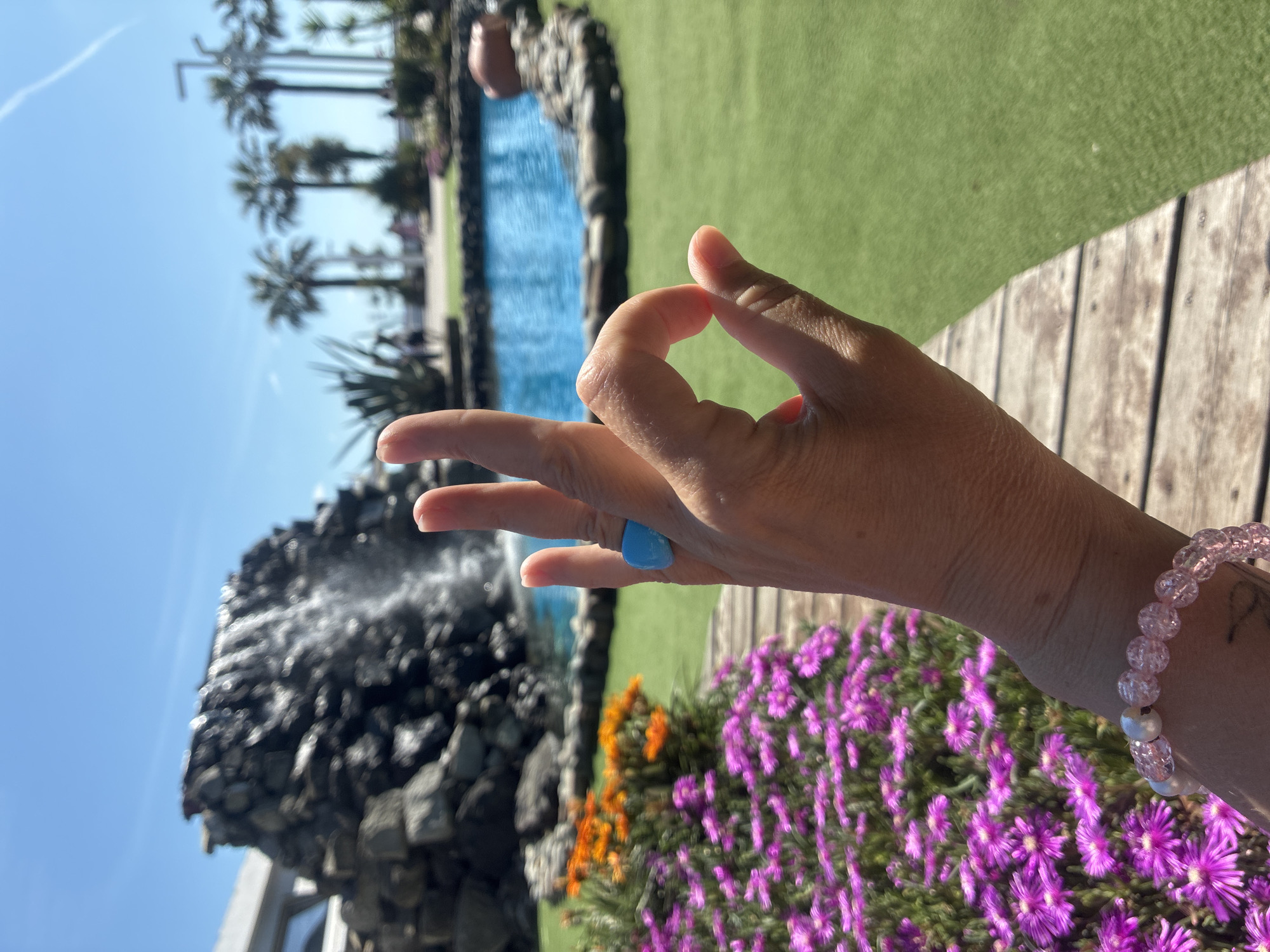 A hand, wearing a blue ring and a pink beaded bracelet, forms an "OK" gesture in the foreground. Behind the hand, a man-made waterfall cascades down a rocky structure, with a blue pool of water situated to its right. The background features a bright blue sky with scattered clouds, palm trees, and a grassy area. Purple and orange flowers are visible to the left of the hand. This scene is located in Sochi, Russia, an area known for its subtropical climate and resort atmosphere.