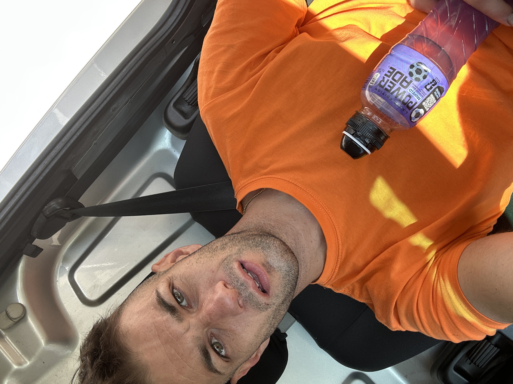 A man is shown from the chest up, sitting in a vehicle, holding a bottle of Powerade. He is wearing an orange t-shirt and has light brown hair and green eyes. The Powerade bottle is purple and partially full. The background is the interior of the vehicle, with a grey metal panel and black upholstery. The lighting suggests it is daytime, with sunlight casting shadows on the man's shirt. The image is a selfie taken by the man. There are no discernible indicators of the specific location within Piperevo, Bulgaria.