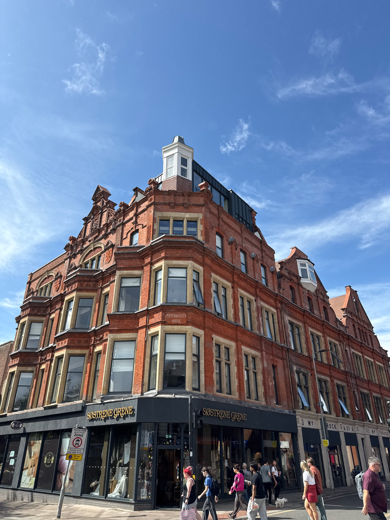 A low-angle, full shot captures the upper floors of a red-brick Victorian building in Exeter, United Kingdom, under a bright blue sky with scattered white clouds. The building features ornate architectural details, including decorative stonework, gables, and bay windows. On the ground floor, a Søstre Grene store is visible, with its name prominently displayed. Several pedestrians are walking on the sidewalk and crossing the street. Some are walking individually, while others are in small groups. One person is walking a small white dog. Traffic signs are also visible, including a pedestrian zone sign. The overall scene depicts a bustling urban street on a sunny day.