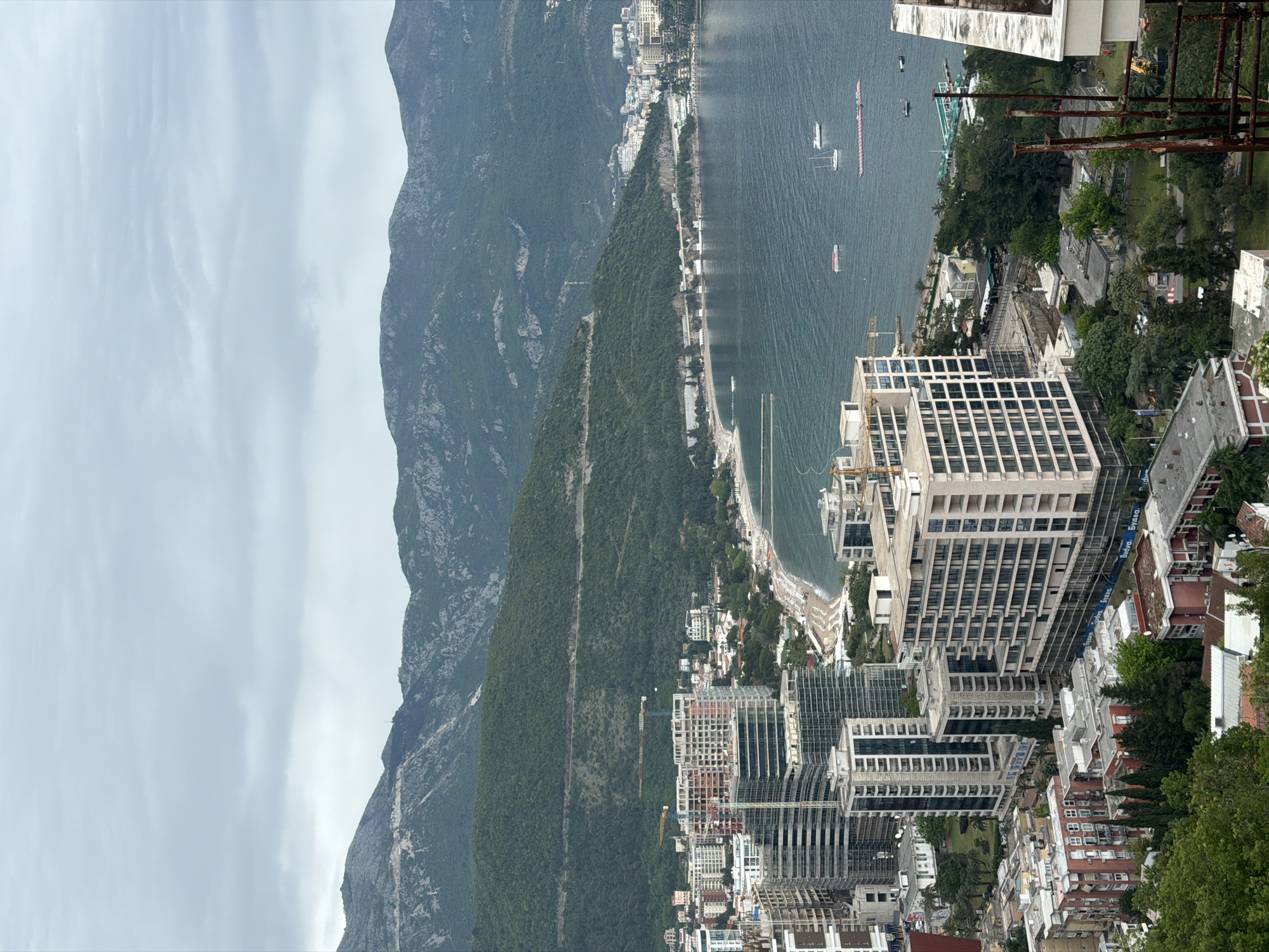 The image shows a high-angle view over Budva, Montenegro, with a focus on the coastal area and surrounding mountains. The foreground and midground contain densely packed buildings, including high-rise residential and commercial structures. Construction cranes are visible amidst the buildings, indicating ongoing development. To the left of the frame, a steep, forested mountain rises sharply. The sea stretches out to the horizon, with boats and other watercraft visible. The sky is overcast with grey clouds.