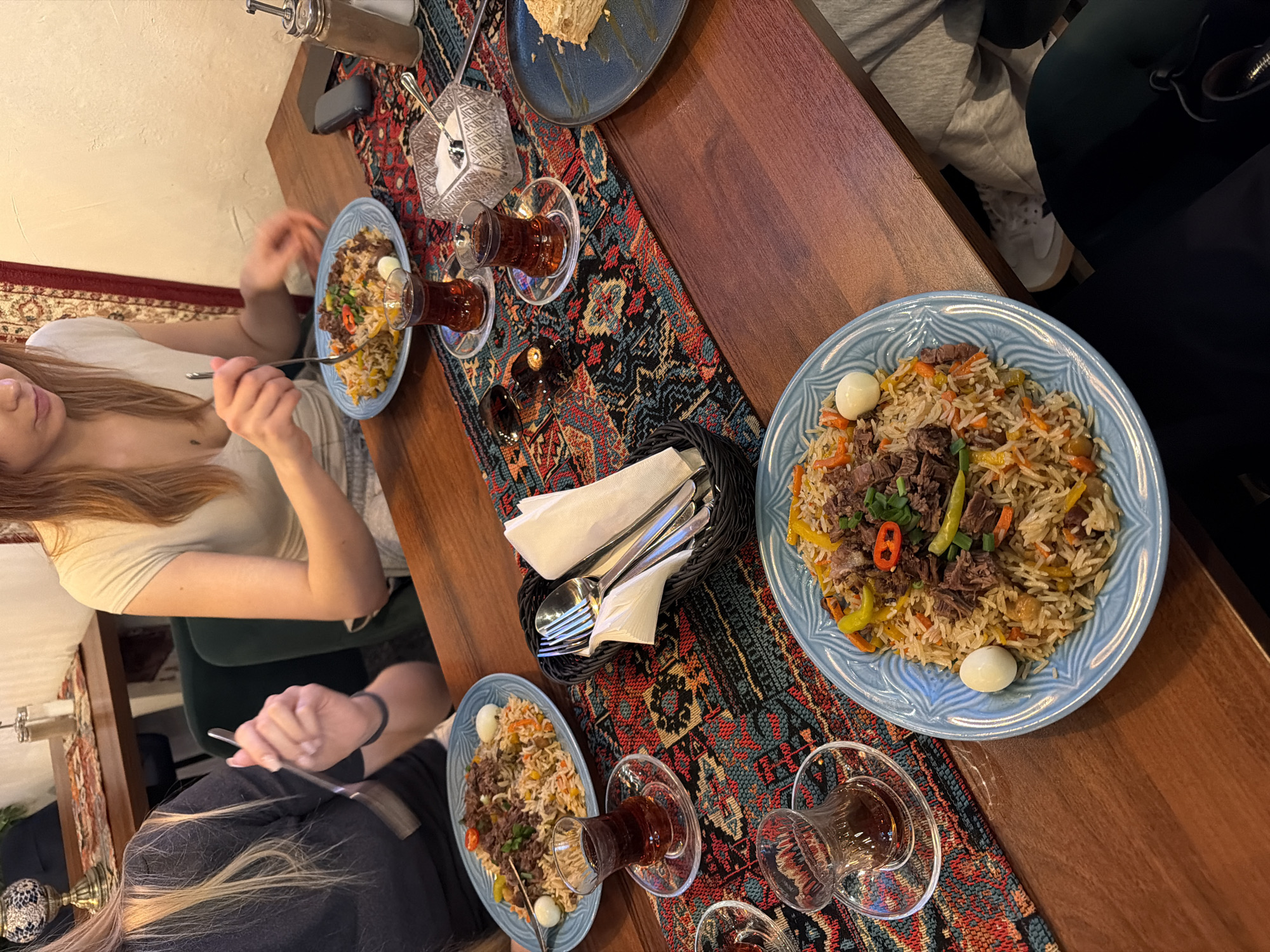 A table in a restaurant setting is shown from an overhead perspective. Three people are seated at the table, two of whom are partially visible. The table is covered with a colorful rug runner with intricate designs. Three blue plates containing a rice dish with meat, vegetables, and what appear to be quail eggs are on the table. Near each plate is a traditional, handleless, tea glass filled with a dark liquid. A basket containing cutlery wrapped in napkins is centered on the table. The table itself is wooden. The background shows the interior of the restaurant with additional patrons, a wall, and a window. The photo appears to have been taken in Warsaw, Poland.