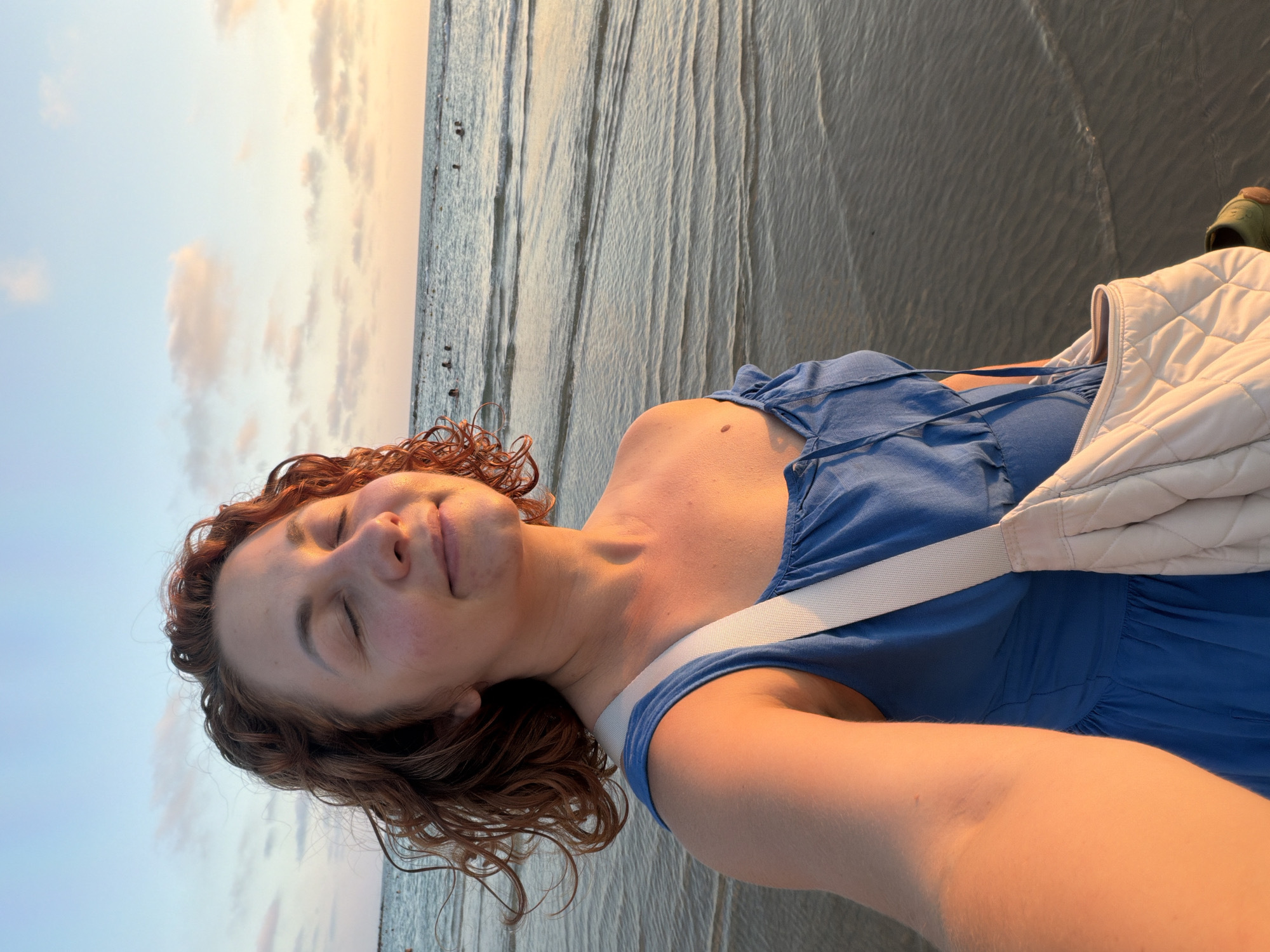 A medium shot captures a woman standing on a beach in Bat Yam, Israel, with her eyes closed and a serene expression. She has reddish-brown curly hair and is wearing a blue dress. A light-colored quilted bag is slung over her right shoulder. The golden light of sunset illuminates her face and shoulder, casting a warm glow. The ocean stretches out behind her, with gentle waves lapping at the shore and a few people visible in the water in the distance. The sky above is a blend of soft blues and pinks from the setting sun, with scattered clouds. The sand beneath her feet is visible in the foreground. The overall atmosphere is peaceful and tranquil.