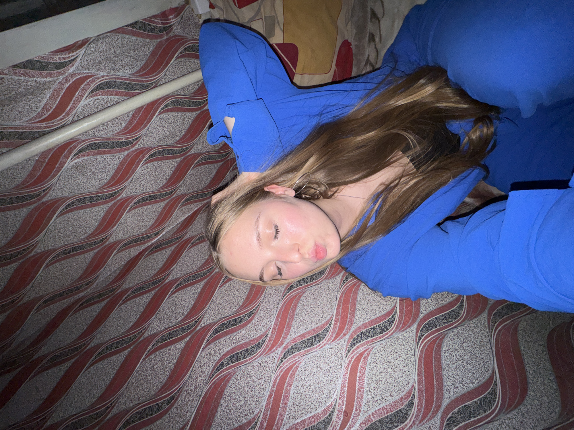 The image depicts a young Caucasian woman with long blonde hair, wearing a blue outfit and a black top. She is positioned indoors, against a backdrop of red and grey wavy patterned wallpaper. Her eyes are closed, and her lips are pursed in a kiss. Her right arm is raised, with her hand resting on her head. The lighting is dim, suggesting an indoor setting at night or in low light conditions. The scene appears to be a bedroom or a similar domestic environment. There are no discernible elements that would indicate a specific location within the city of Perm, Russia.