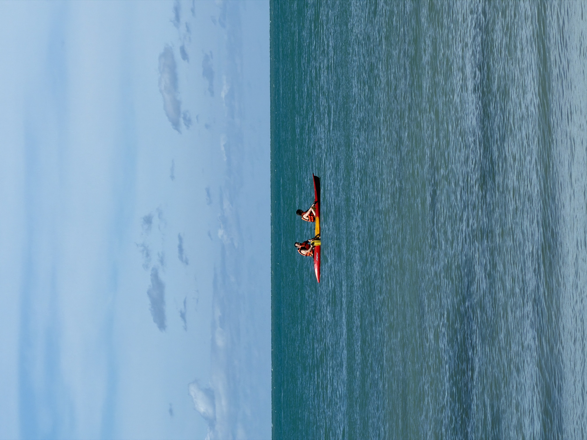 This media file shows two people in a red kayak on a calm, blue ocean. The image is oriented vertically, with the sky occupying the left half and the ocean the right half. The sky is a pale blue with scattered white clouds. The ocean has gentle ripples, indicating a peaceful setting.
The two individuals in the kayak are both wearing life vests. One person is positioned at the front of the kayak, and the other is at the back. They appear to be paddling, engaged in recreational kayaking. The kayak itself is a vibrant red.
The scene suggests a clear, bright day, likely during daylight hours, given the illumination on the water and sky. The calm nature of the sea and the presence of kayakers point towards a pleasant weather day suitable for water activities. The image evokes a sense of tranquility and enjoyment of the outdoors.
No visible text is present in the media file. The location context is Baan Plai Laem, Thailand, which suggests a tropical or subtropical coastal environment.