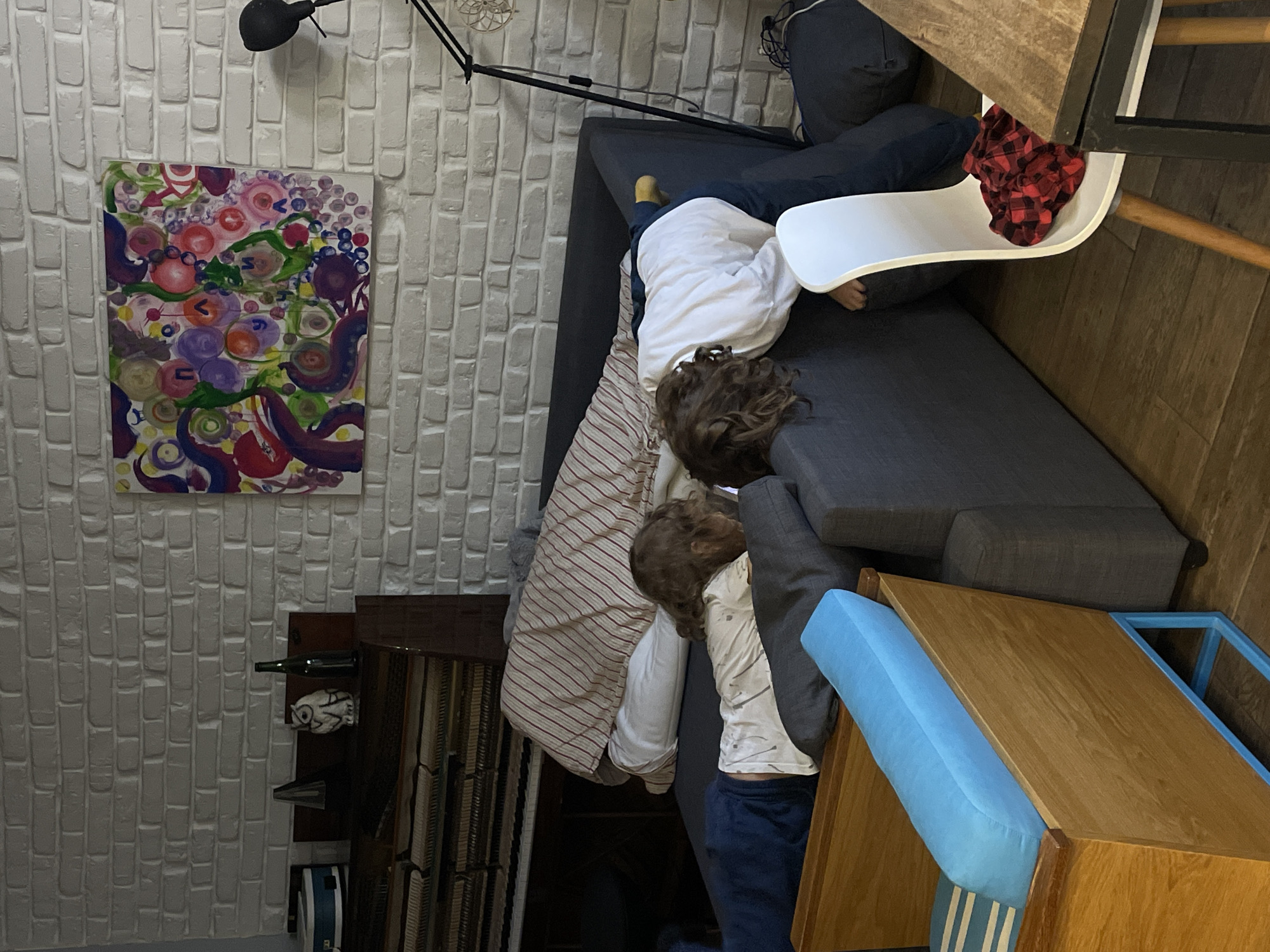 A wide-angle, rotated, high-angle shot shows a room with a white brick wall on the left. On the wall is an abstract painting featuring circular shapes in pink, red, purple, and green. A black adjustable lamp is positioned above the painting. Next to the wall is a dark wood cabinet holding framed artwork and a small decorative owl figurine, along with a bottle of wine.
Two children are on a gray couch covered with a striped blanket. One child is wearing a white top and dark pants, partly obscured by the couch. The second child, wearing a white shirt, is reclining on the couch. A white high chair with a red and black checkered pattern rests near the children and the couch.
A wooden cabinet with a light blue cushion is in the lower right corner. The floor is wood-paneled.
There is no information to confirm this is in the city of Moscow, Russia.