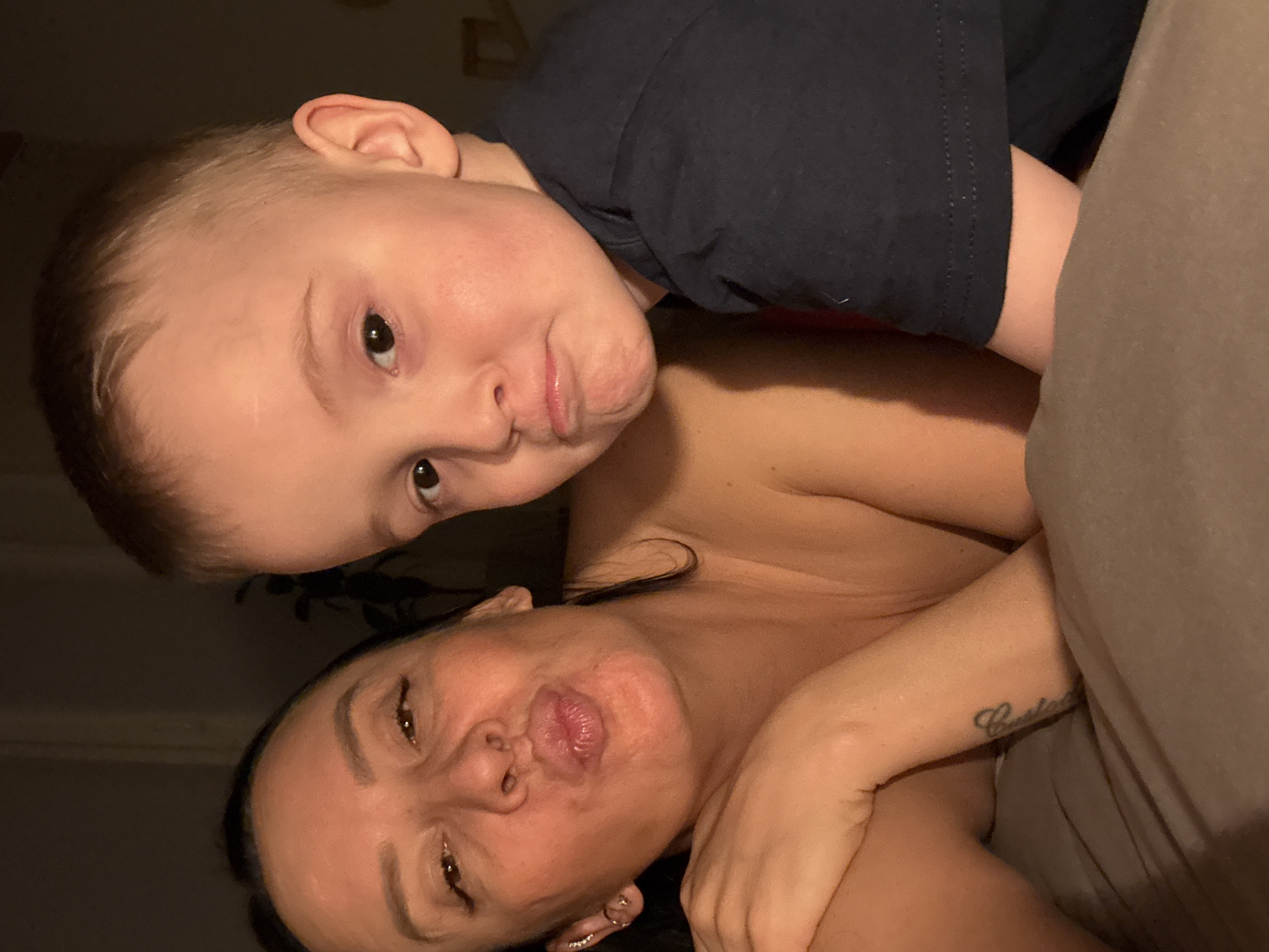 The image captures a close-up, intimate moment between an adult female and a young child, likely indoors during the evening or night due to the dim lighting. The scene appears to be set in a bedroom, with a bed or blanket visible. The provided context indicates the location is Chaikovsky, Russia, though no specific visual cues in the image confirm this.
The subjects are a young child, appearing to be a boy, and an adult woman. The child is positioned above the woman, resting on or near her. He wears a dark blue t-shirt and looks directly at the camera with a playful, pouting expression, his lips slightly pursed.
The woman is lying down beneath the child, also looking towards the camera. She has dark hair and is shirtless, partially covered by a light brown or beige blanket. She mirrors the child's playful expression, pursing her lips. Notable details on the woman include a small, embellished ear piercing on her left earlobe and a tattoo on her right forearm. The tattoo is script text, partially visible, and appears to begin with "Christa" or a similar word.
The activity taking place is a shared, casual moment, possibly a selfie, with both subjects engaging playfully with the camera through their pouting expressions. The emotions conveyed are warmth, affection, and playfulness, highlighting a strong bond between the two individuals.