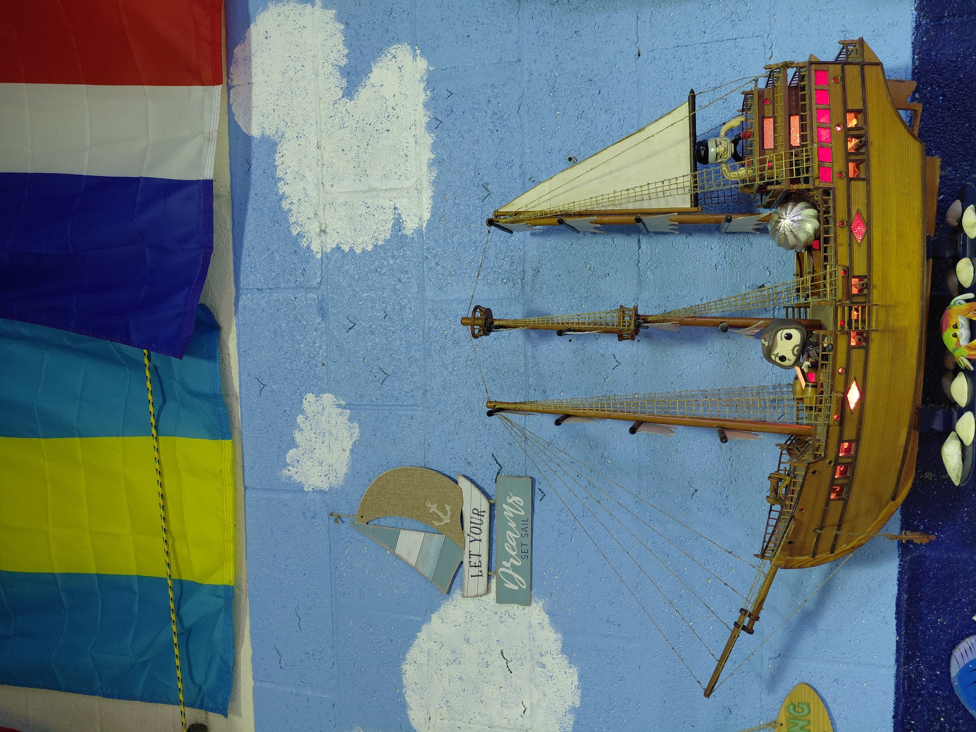 The image shows a wall in what appears to be a nautical-themed room in Cutler Bay, Florida, United States. The wall is painted blue with white cloud-like shapes. A wooden model ship is mounted on the wall, oriented vertically. The ship has multiple masts and sails, with small figures positioned on the decks and rigging. The ship's interior is illuminated with red lights. To the left of the ship, several flags are hung on the wall, including the flag of the Netherlands, and what appears to be the flag of Buenos Aires (Argentina). Below the ship is a decorative sign with a small sailboat and the words "LET YOUR Dreams SET SAIL." Other decorative items and nautical motifs are visible around the room.