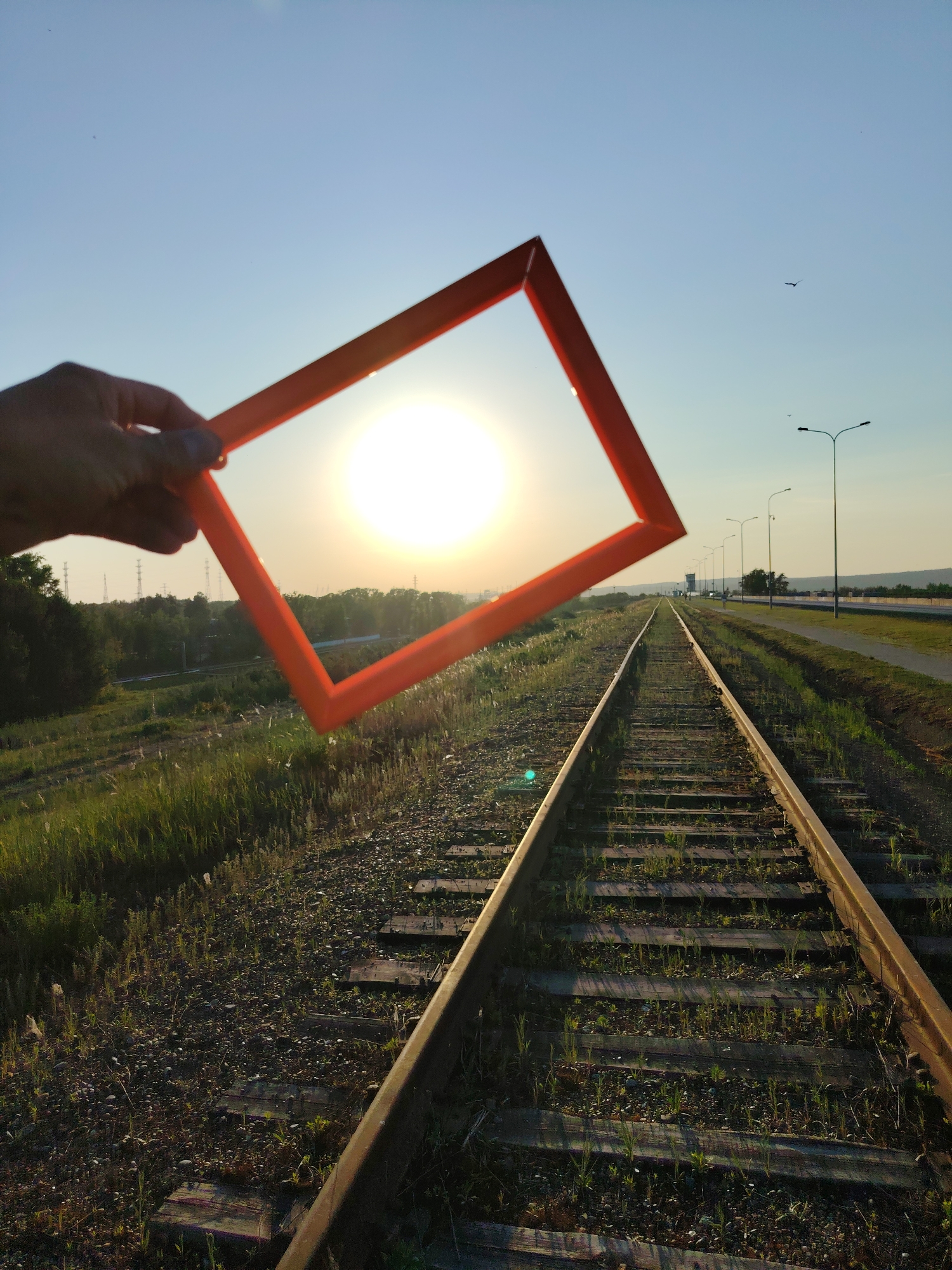 A hand holds an orange square frame up to the sun, framing the sun in the center of the frame. In the foreground, there is an overgrown train track leading towards the sun and a road parallel with street lamps. The location is a railway track in Chaikovsky, Russia.