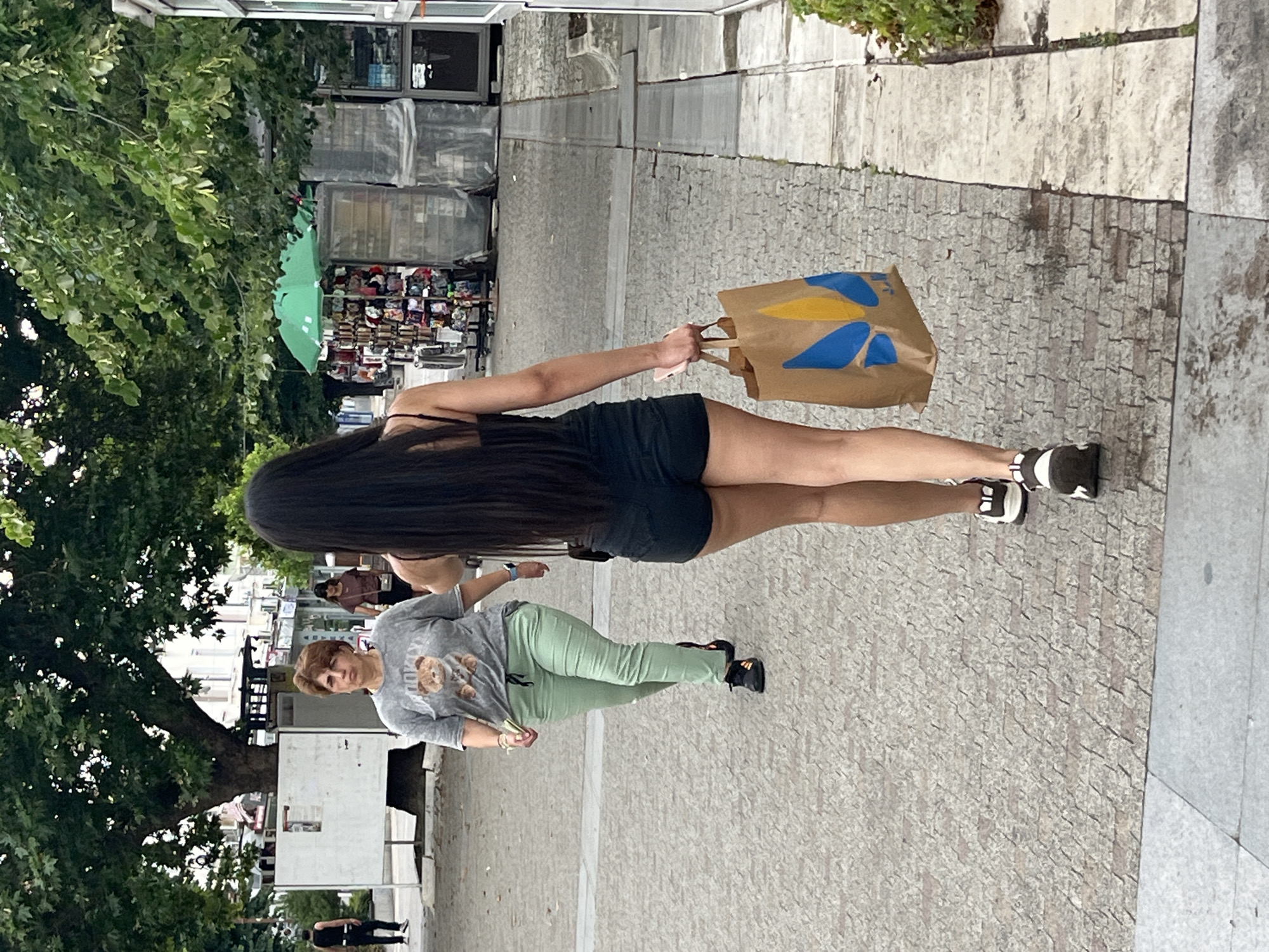 In Sliven, Bulgaria, on a pedestrian walkway with cobblestone paving, a woman with long dark hair, wearing a black tank top and black shorts, walks away from the camera carrying a brown paper shopping bag with a blue and yellow design. She is wearing white and black sneakers. To her left and slightly behind her, another woman with short brown hair, wearing a grey t-shirt with a bear graphic and light green pants, walks in the same direction. She is wearing black sneakers. In the background, trees, buildings, and a market stall with a green umbrella are visible.