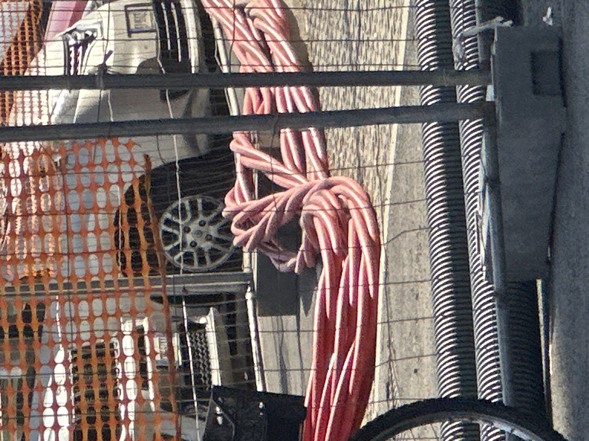The image, likely taken in Milan, Italy, shows a construction or maintenance site. A safety barrier, composed of orange plastic mesh, is partially visible, behind a grey metal construction fence with black plastic screening. The site contains equipment and materials. A stack of red and white striped hoses is in the middle ground. To the right of the hoses, two runs of ribbed black flexible tubing are stacked beside a light grey concrete support. In the background, a light-colored vehicle with a multi-spoke wheel is partially visible. The bottom portion of a bicycle tire is visible in the foreground. The scene is observed in daylight conditions.