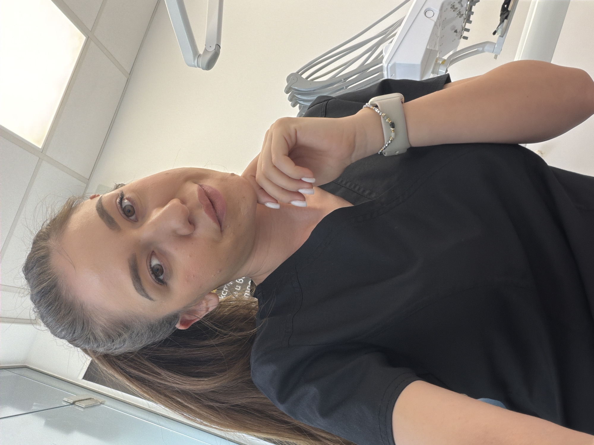 The image shows a young woman wearing black scrubs, suggesting she might be a medical professional, possibly in a dental or medical office given the equipment visible in the background. She has long brown hair pulled back and is wearing makeup. A silver smartwatch is on her left wrist. The scene appears to be indoors, in a well-lit room with a tiled ceiling and what looks like dental equipment or a medical light fixture overhead. The woman is looking directly at the camera with a neutral expression. There is no visible text in the image. The image is taken from a slightly low angle and is oriented sideways.