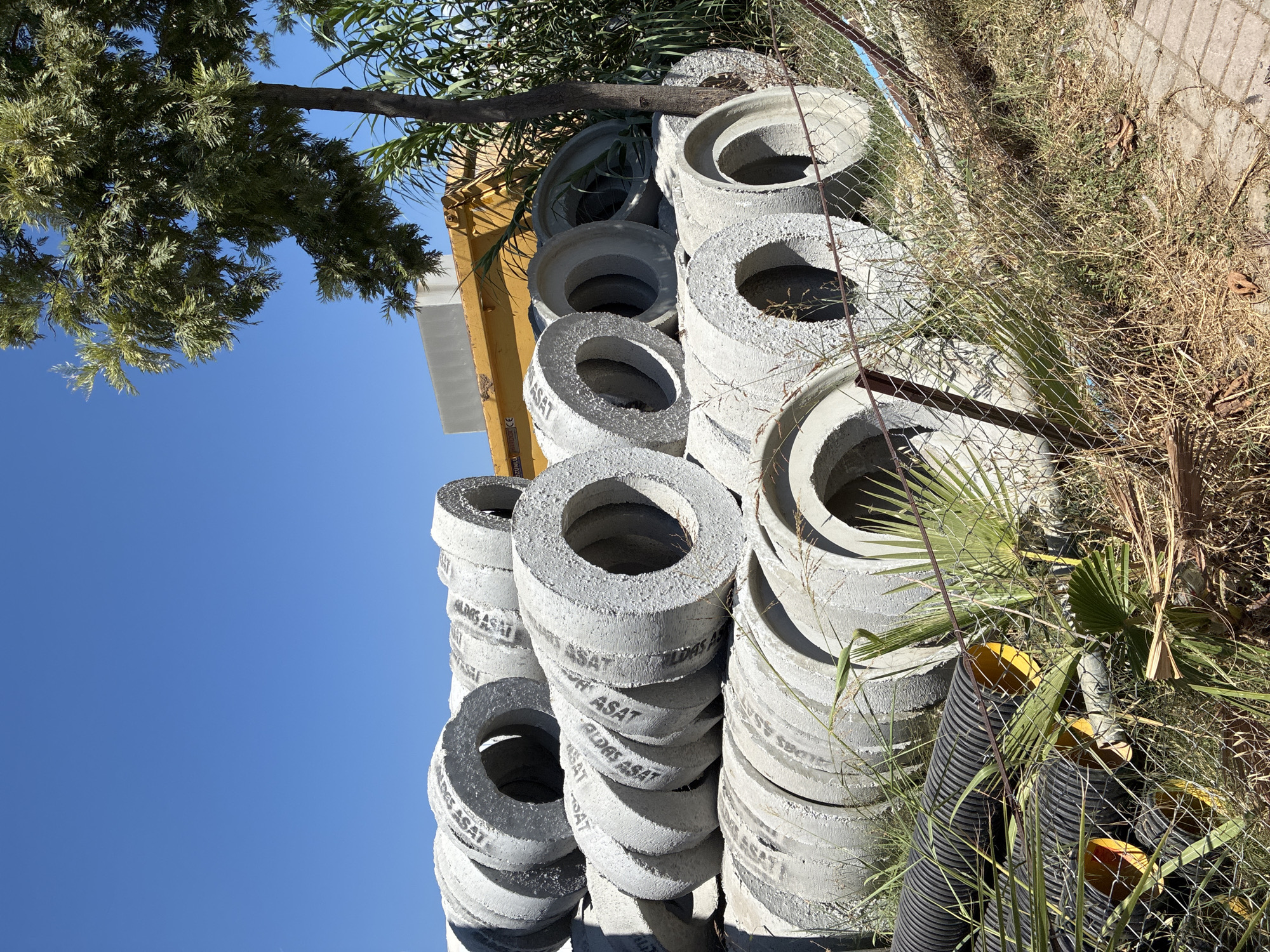 A stack of concrete rings, likely for sewage or water systems, sits piled outdoors in a yard in Alanya, Turkey. The rings are gray, weathered, and have "ASAT" imprinted on them, which stands for "Alanya Altyapı ve Kanalizasyon Müdürlüğü" or Alanya Infrastructure and Wastewater Directorate. In the background, a yellow construction dumpster is visible, along with tall green plants and a tree with feathery leaves. A chain-link fence with some dry grass and a fallen palm frond lies in the foreground. The sky is a clear, bright blue.
