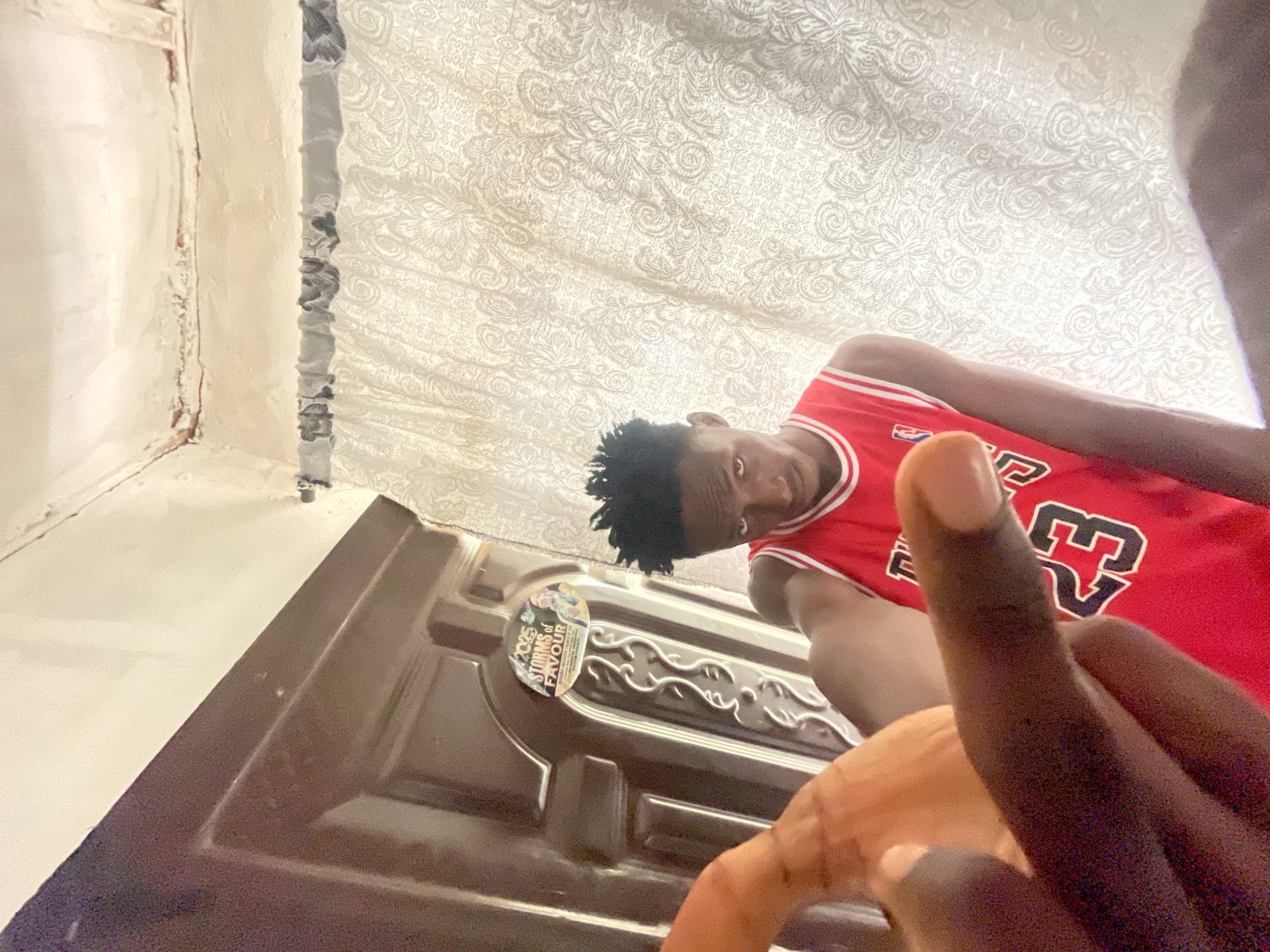 A low-angle shot shows a person in a red basketball jersey with the number 23, possibly wearing a Chicago Bulls jersey, looking down at the camera. The person has short black hair. The background consists of a patterned white curtain and a dark brown door with a sticker on it. The sticker appears to say "2025 Storms of Favour." Two fingers are in the foreground. The photo appears to be taken indoors. There is no definitive way to confirm the location is Lokoja, Nigeria, from the image alone.