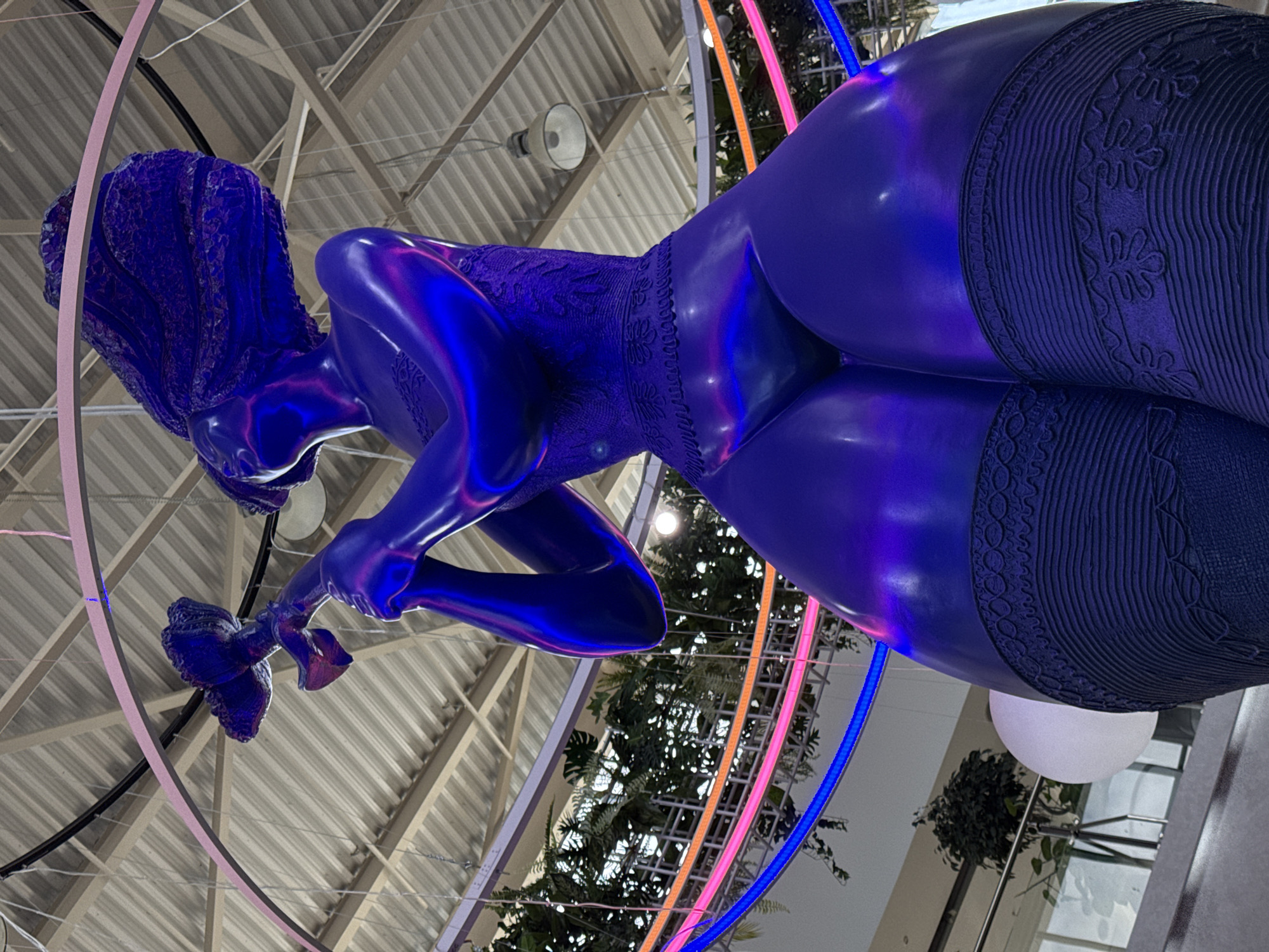 A low-angle shot captures a large, shiny, indigo-colored sculpture of a reclining woman. The figure wears a patterned garment covering her torso and high-patterned stockings that reach her upper thighs. Her head is tilted downward, and she holds an indistinct object in her hand. The sculpture is indoors, suspended within a circular frame of light blue, pink, orange, and black bands. The backdrop consists of a corrugated metal ceiling, hanging light fixtures, indoor greenery, and a white globe lamp in the background. This sculpture is located in the Nizhny Novgorod, Russia airport.