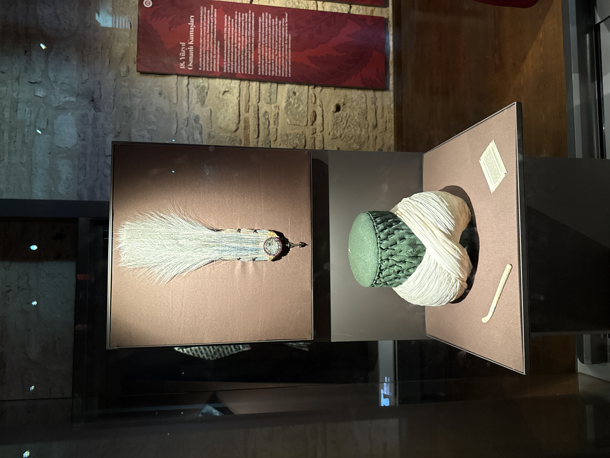 In an indoor museum exhibit in Istanbul, Turkey, a green fez-like hat with white cloth wrapping is displayed on a pedestal. Above the hat, a large framed display showcases a decorative ornament featuring a cluster of white feathers and intricate metalwork with colored gems. To the right, a red informational placard with Turkish text is mounted on a stone wall. A small, bone-colored object, possibly a tool or decorative item, rests on the pedestal in front of the hat, alongside a small, rectangular card with text. The background features rough stone walls and wooden flooring, suggesting a historical setting within the city.