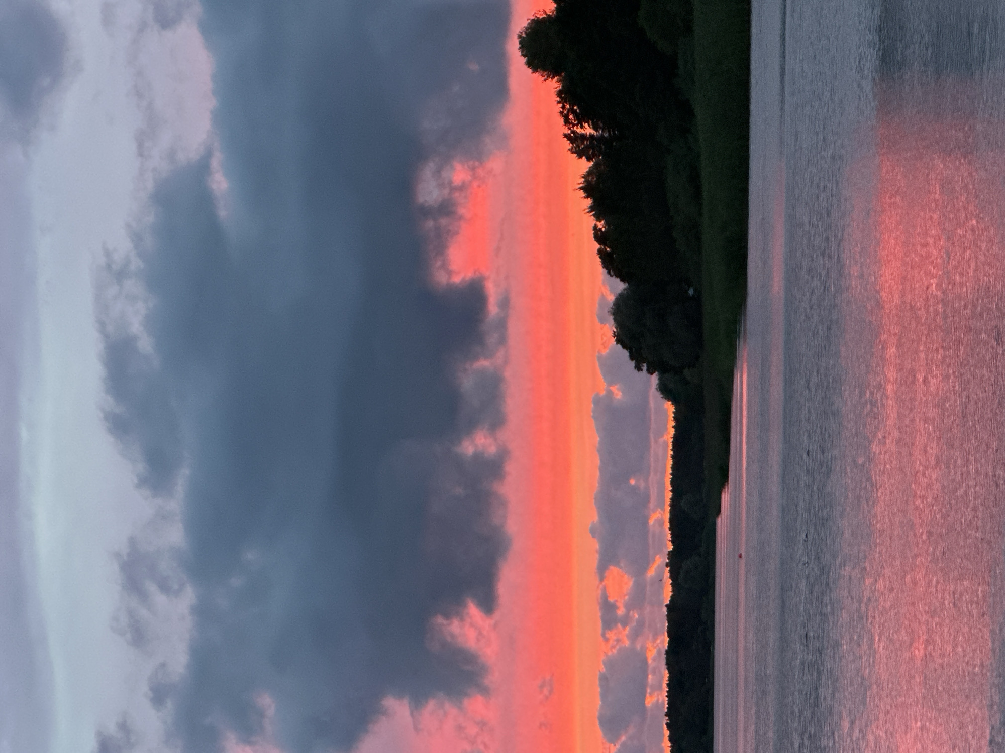 The image depicts a body of water, likely a lake or a bay, with a dense treeline along the distant shore. The sky is dramatic, filled with dark, heavy clouds near the top and vibrant pink and orange hues lower down, suggesting a sunset. The water reflects the colorful sky, creating a shimmering pink surface. The overall atmosphere is serene and natural, with no human presence or urban elements visible. The description of the location as being in Tallinn, Estonia, cannot be verified from the visual information provided in the image itself.