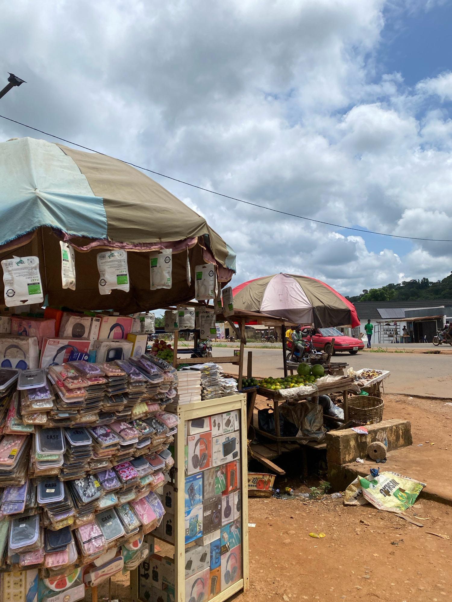 A street market stall in Iworoko, Nigeria, displays an assortment of mobile phone accessories, including phone cases and earphones, under a large tent. A second, smaller stall to the right features fruit and other produce. In the background, a red car and several motorcycles are visible, along with pedestrians and buildings. The scene is set on a dirt road under a cloudy sky.