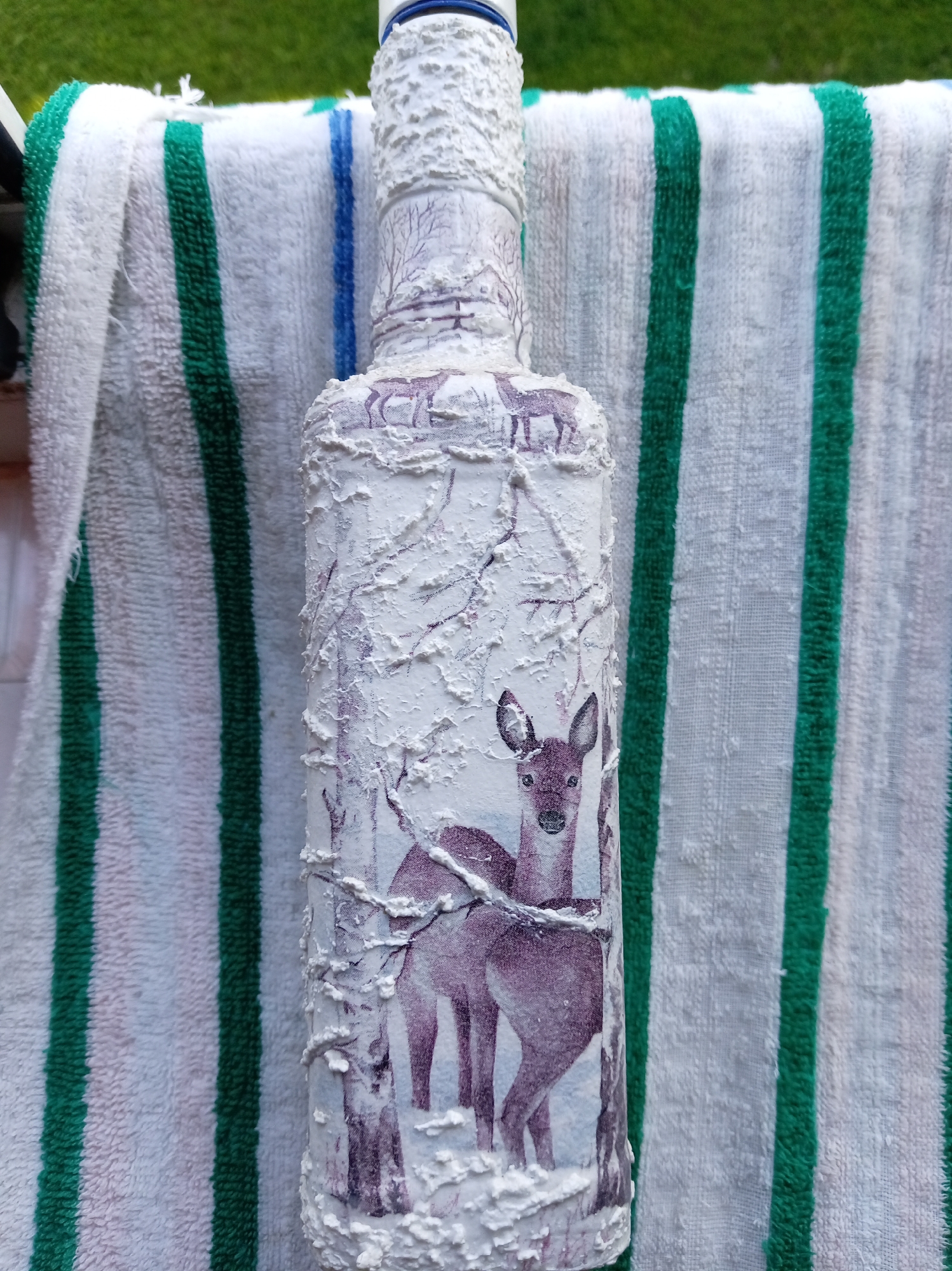 A white, textured bottle adorned with a winter scene featuring deer and bare trees is placed on a striped towel. The towel has vertical stripes in white, green, and blue. The background is out of focus and appears to be green grass, suggesting an outdoor setting. The specific location in Бараўлянскі сельскі Савет, Belarus cannot be determined from the image alone.