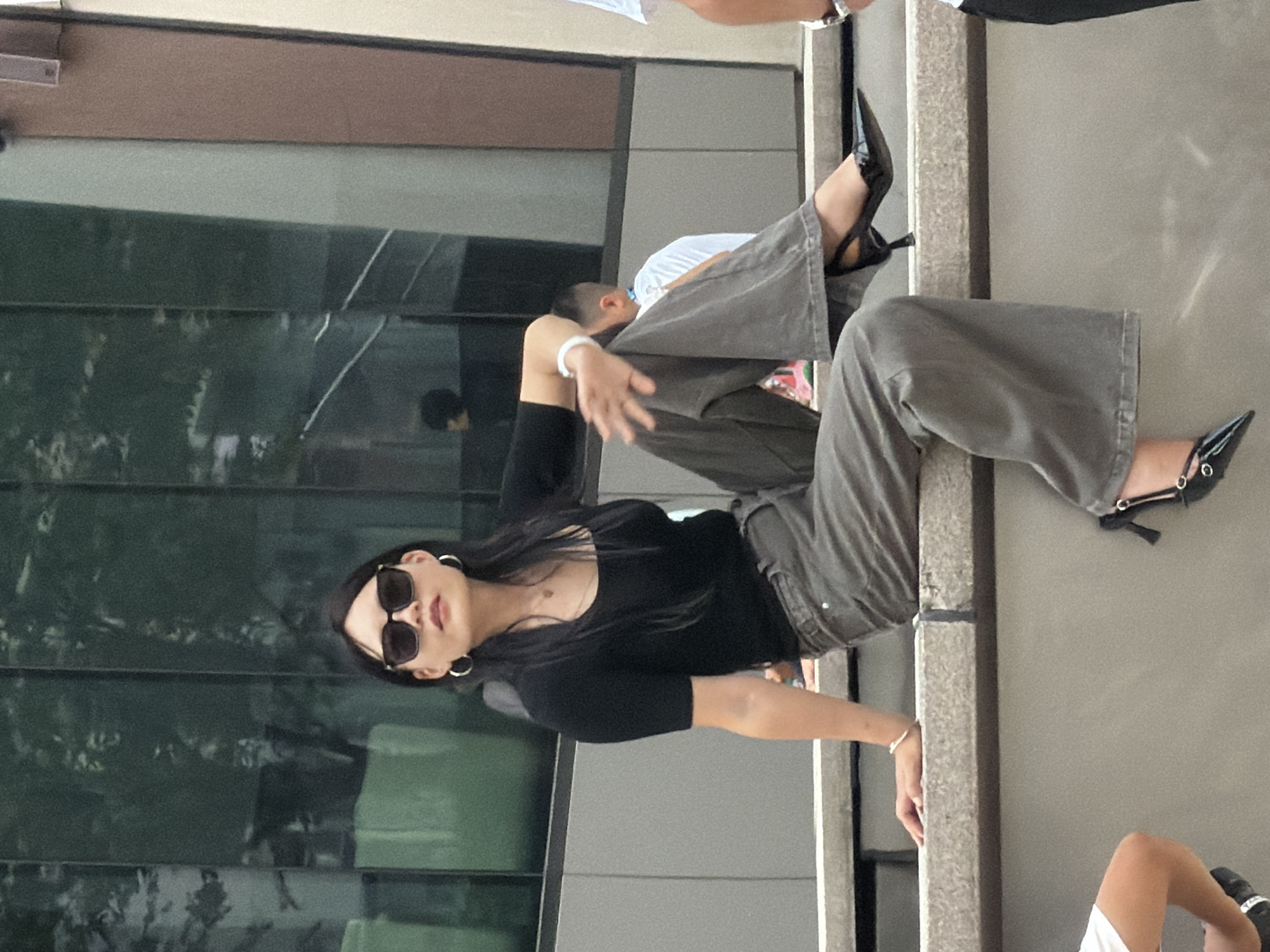 A woman with long dark hair and sunglasses is sitting on a concrete ledge in an urban environment, with large reflective windows behind her. She is wearing a black short-sleeved top and wide-leg grey pants. On her feet, she has black pointed-toe heels with ankle straps. She is wearing large hoop earrings and a bracelet on her left wrist. Her right hand is resting on her thigh, and her left hand is extended slightly. The background shows reflections of trees and a building structure. Other people are visible in the background, out of focus. The scene appears to be in a public space, possibly near a modern building in Yuzhong District, China.