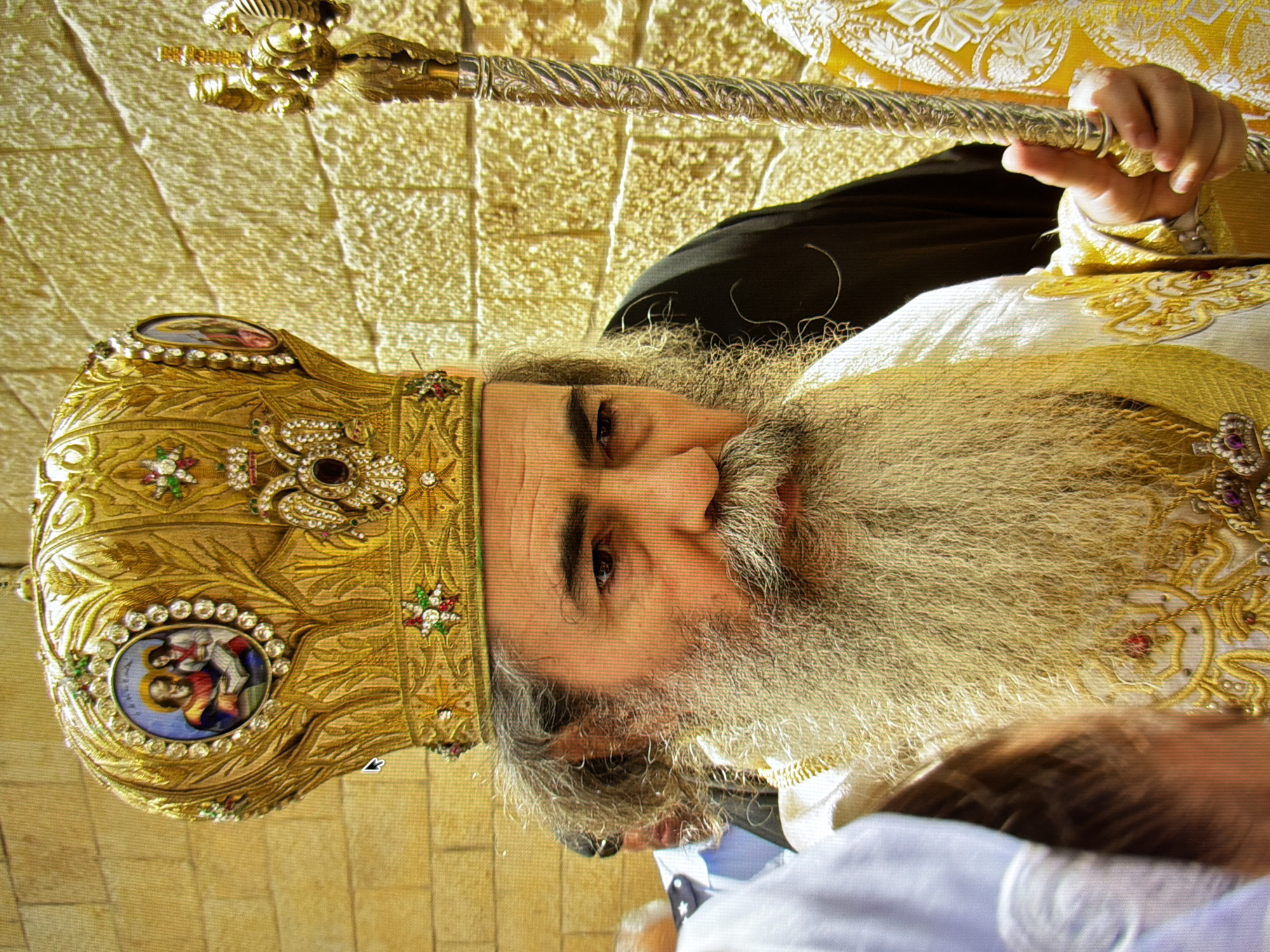 A medium shot captures a man wearing a golden, jeweled mitre and ornate white and gold religious vestments. He has a long, light-colored beard and is holding a golden staff. He is standing in front of a light brown brick wall. There are other individuals partially visible in the background, some wearing white and blue. The man's expression is neutral, and he is looking slightly to his right. The scene appears to be outdoors.