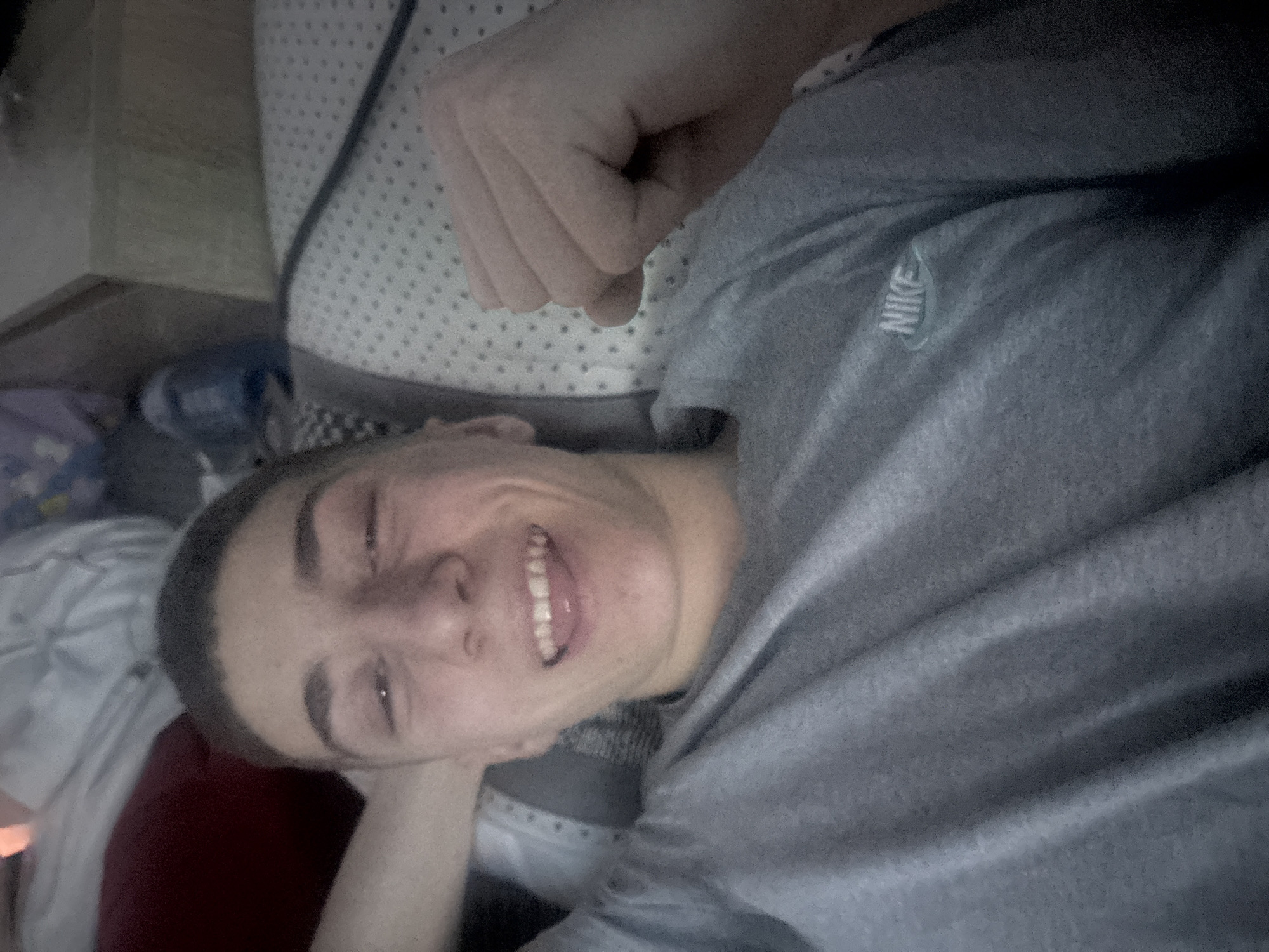 A person is lying down in what appears to be a bed. The person is wearing a gray Nike t-shirt. Their head is resting on a hand, and they are smiling. There is a bedspread with a pattern visible, and other soft objects are visible around the person. The location is likely an indoor setting. The city is Gorni Bogrov, Bulgaria.