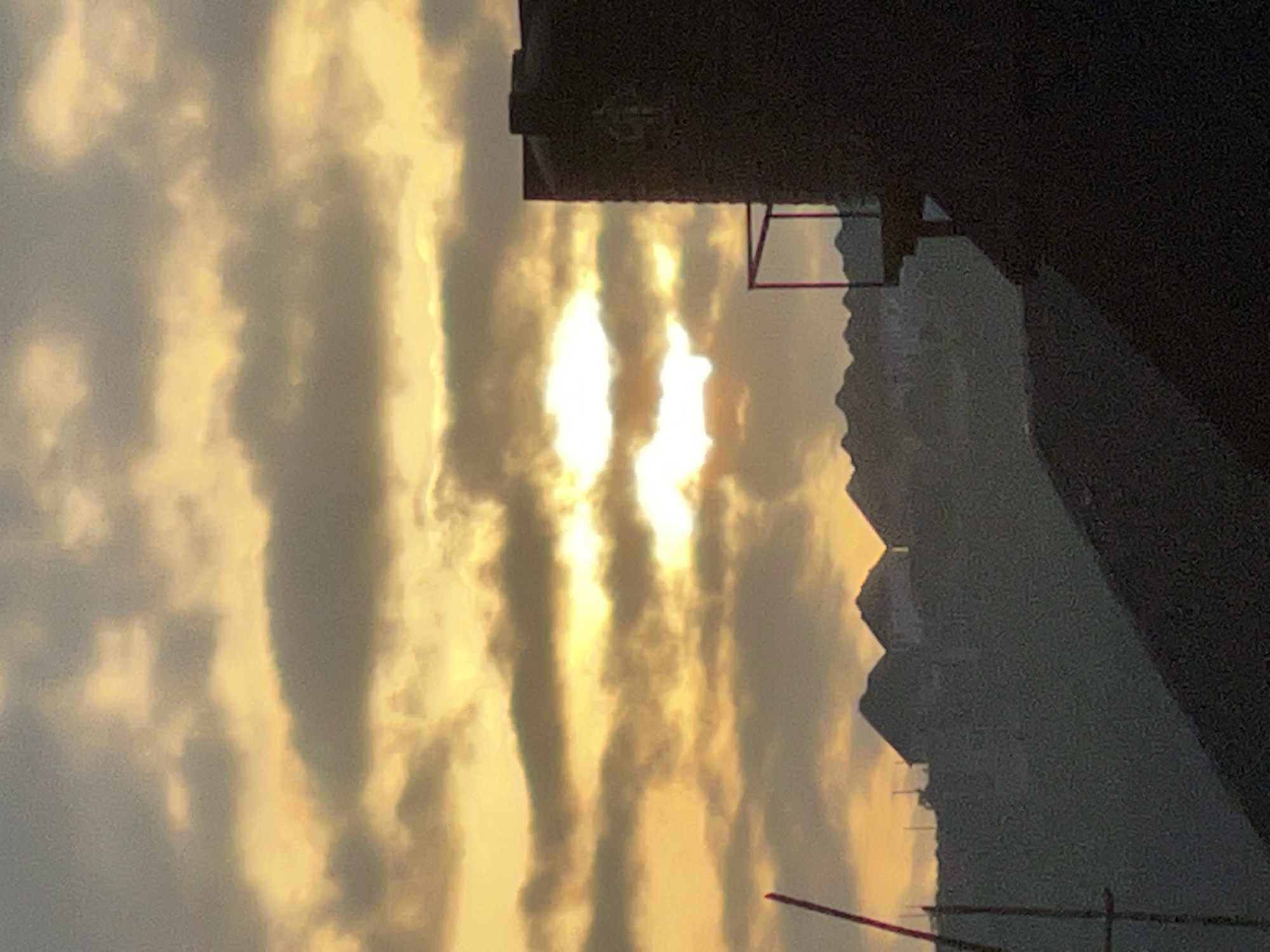 The image depicts a silhouetted view of houses on a hill against a cloudy, golden sunset sky. The sun is partially obscured by the clouds, creating a bright glow. In the foreground, a dark, textured structure, possibly a water tank, rises on the right side of the frame, with a metal railing attached. To the left, a red pole and scaffolding are visible. The setting appears to be an elevated area overlooking a residential neighborhood. Given the architectural style of the houses and the presence of such infrastructure, this scene could be located in a developing or established residential area within the city of Abuja, Nigeria, such as Maitama Hills or Apo Resettlement.