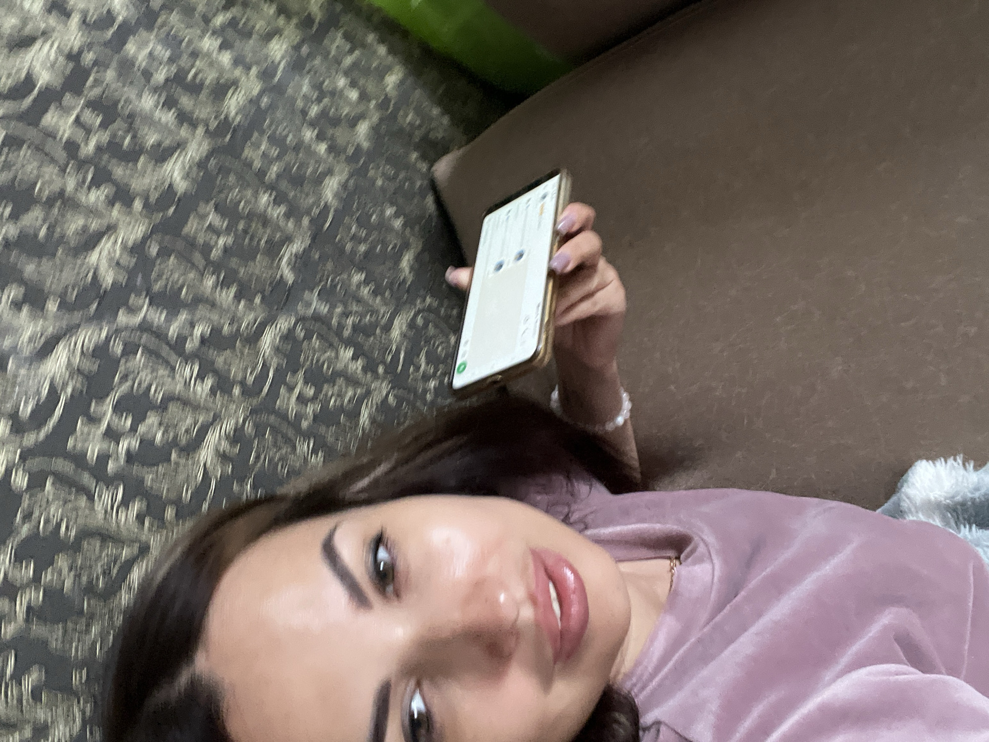 A woman is holding a smartphone in her right hand, looking at the camera. The smartphone displays a messaging application. She is wearing a pink velvet shirt and a pearl bracelet on her right wrist. The background consists of a patterned wallpaper and a brown couch. A green bottle is partially visible on the right side of the image.