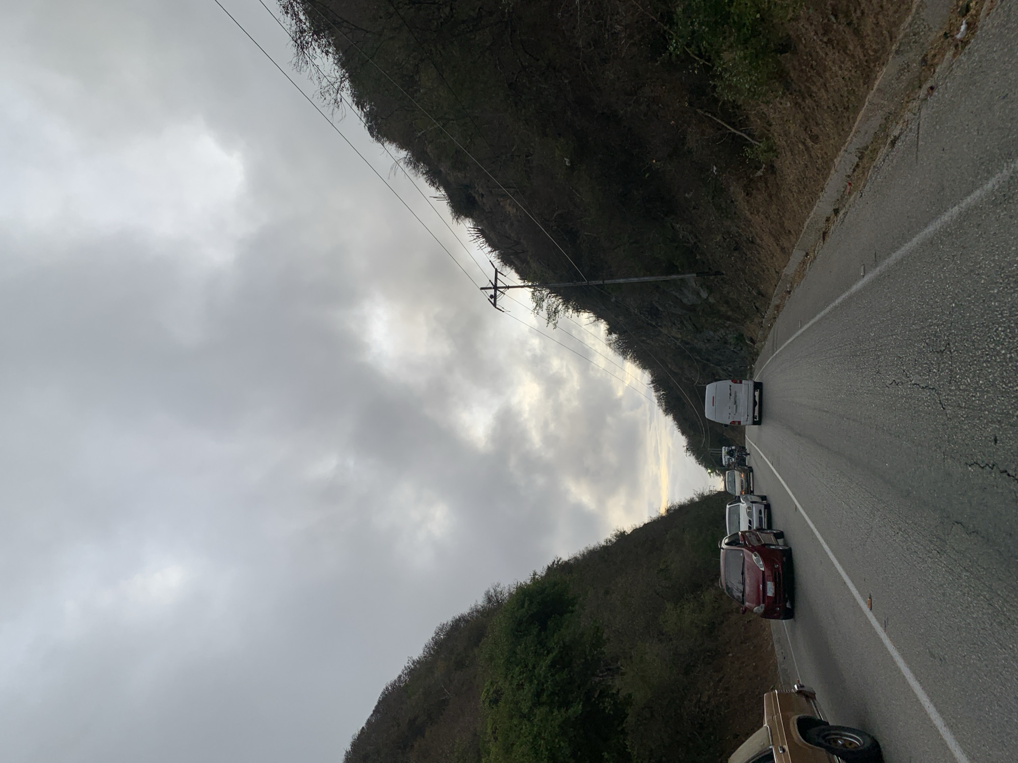 The image shows a road in La Asuncion, Venezuela, with a line of vehicles. The sky is overcast. Mountains rise on both sides of the road. Power lines run along one side of the road.