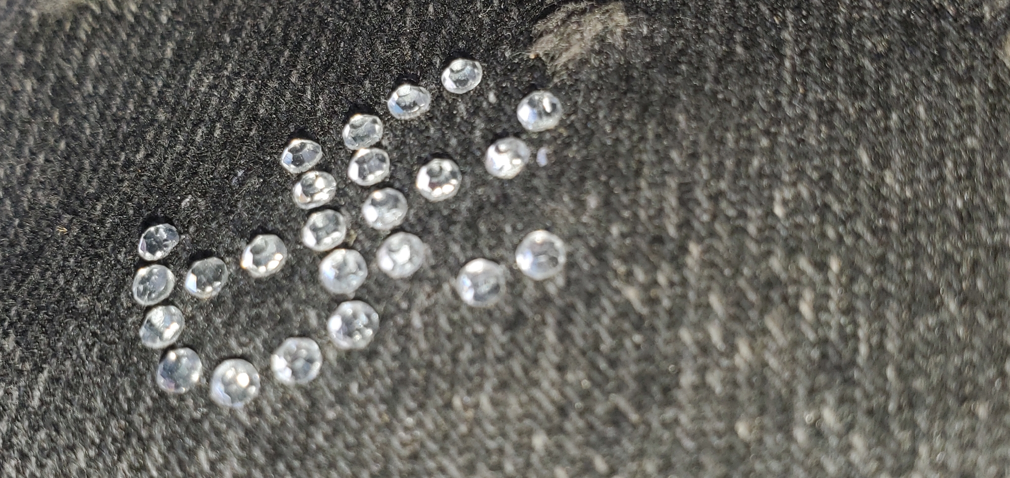 The image displays a collection of small, clear, faceted stones, likely diamonds, scattered across a dark, textured surface that appears to be fabric. The stones are irregularly spaced and vary slightly in size and shape, with many exhibiting a polished, reflective quality. The lighting casts subtle shadows and highlights on the stones, emphasizing their facets and transparency. The background is uniformly dark and granular, suggesting a close-up view of the stones on a surface like velvet or felt, commonly used for displaying jewelry. The scene is devoid of any human presence, animals, or specific environmental elements that would indicate a particular location within Bukuru, Nigeria, or any other city. The image focuses solely on the presentation of the gemstones.