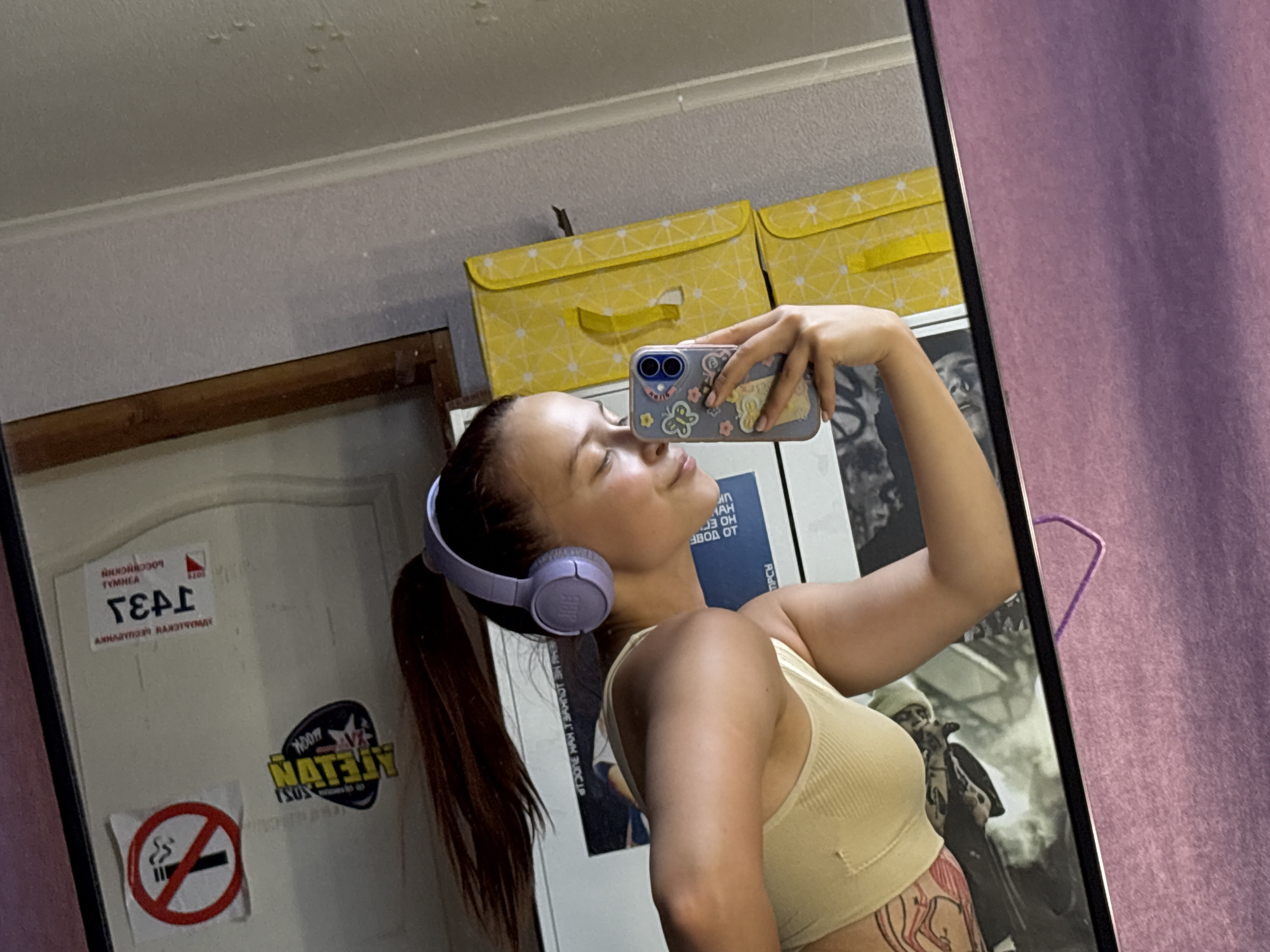 A woman wearing headphones takes a mirror selfie in what appears to be a domestic setting. She is angled towards the mirror, holding a phone with a decorative case. A closed white door is visible in the background with affixed stickers including a no-smoking sign and an upside-down graphic that includes the words "ATELON" and "MOGK". On the upper part of the wall is a piece of paper with "TIPCH" and "43" visible. The text includes "ТИМНЕА," "Боссиискии", "АХНЛОУПОЭI NAXЭТЧҮМДҮ", and "1071". There are yellow storage boxes on top of the door. There is a poster to the right that has a visible person who has a lit cigarette. The woman is wearing a light yellow cropped top with a pattern on her lower abdomen. Behind the mirror, a section of purple curtain is visible. Based on the sticker on the door, the location may be in the city of Прикамский, Russia.