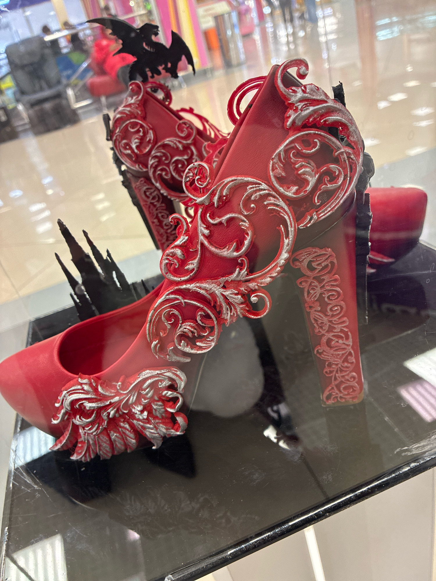 A pair of ornate red high-heeled shoes are displayed on a glass surface. The shoes are decorated with silver filigree designs and appear to have gothic-themed embellishments, including what looks like a bat on the back of one shoe and sharp, dark spikes on the heel of the other. The background is blurred and appears to be an indoor commercial setting, possibly a store, with other displays and reflections visible through a glass barrier. The specific location in Moskovskiy, Russia, is not identifiable from the image alone.