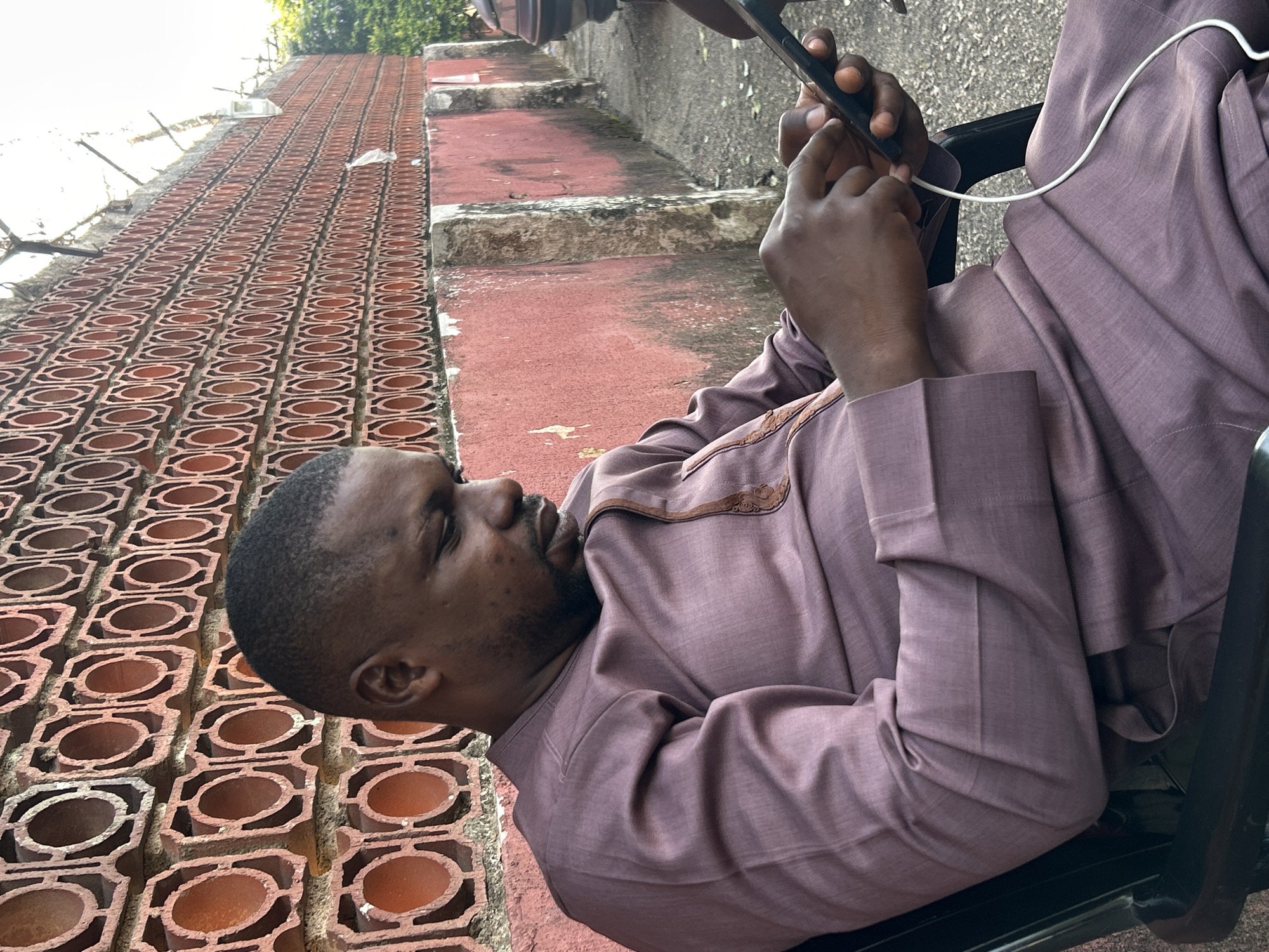 An adult male is seated outdoors in a black chair, engrossed in a black smartphone connected to a white charging cable, which rests on his lap. He is dark-skinned with short hair and a beard, wearing a lavender-colored traditional tunic and trousers. The setting is in Jos, Nigeria. Behind him is a tall wall constructed from reddish-orange, hollow terracotta bricks arranged in a repetitive pattern of interconnected octagonal frames surrounding circular openings. The top of this wall features barbed wire. Beneath the patterned brickwork, a lower section of the wall is painted a weathered reddish-pink. The ground is an uneven grey surface. In the distant right background, green foliage is visible next to the rear portion of a dark-colored vehicle.