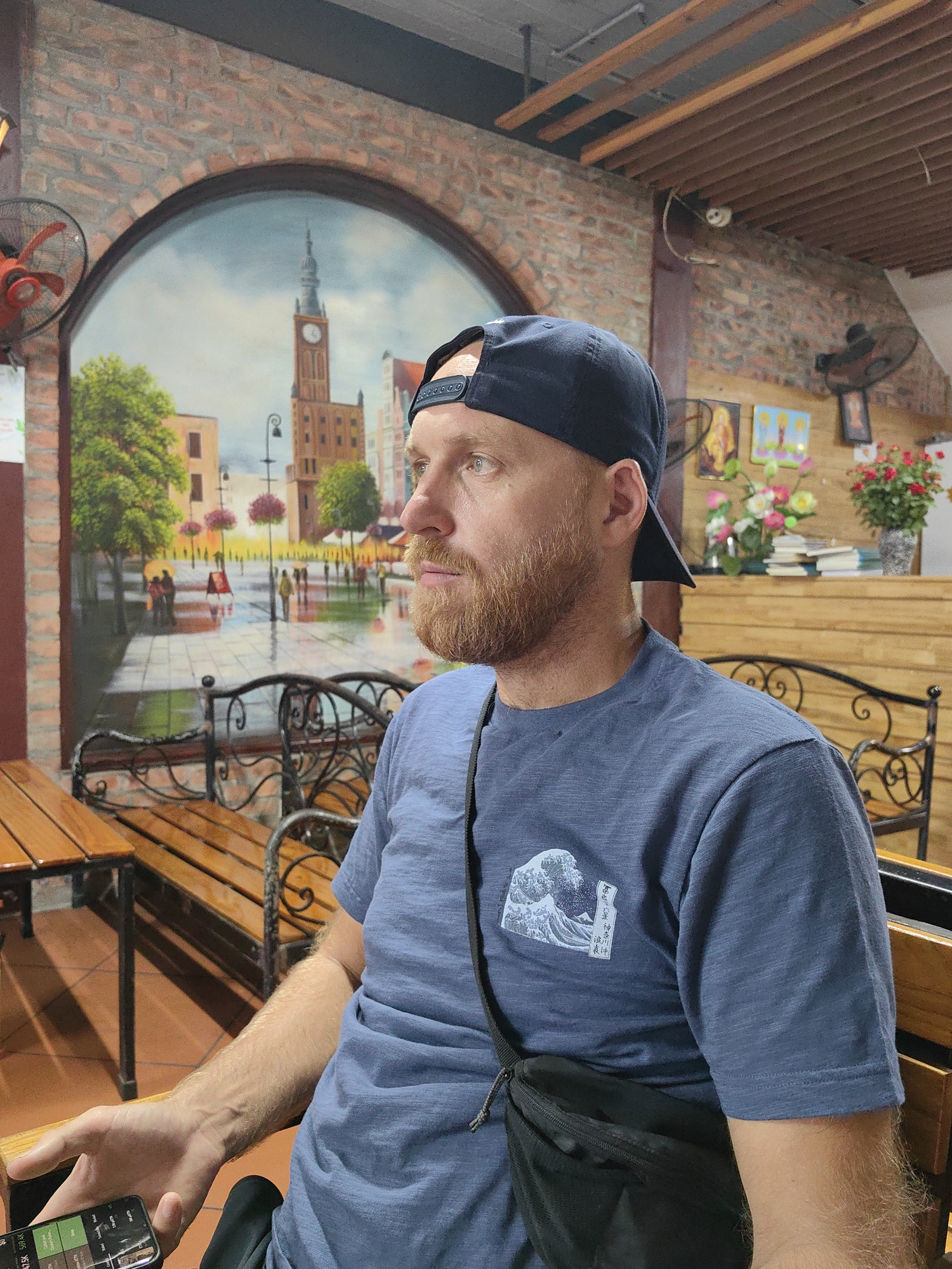 A man with a beard and a navy blue baseball cap is seated in an indoor location, likely a restaurant or café. He's wearing a blue T-shirt with a graphic of a wave and Japanese characters on the upper left chest area. He is holding a smartphone in his left hand. A black shoulder bag is across his chest. The setting appears to be indoors, with wooden benches and tables. Behind him is a painted mural depicting a European city scene, possibly Warsaw or Gdańsk in Poland, featuring a tall clock tower and street with figures. The indoor space is characterized by brick walls, a wooden ceiling, and several fans, including a red fan to the left of the mural and a brown fan on the opposite wall. The scene suggests the location is in Vietnam. Based on the architectural style, including the brick and the style of the mural, it is likely this image was taken in Hà Nội, Vietnam, where such aesthetics can be found.