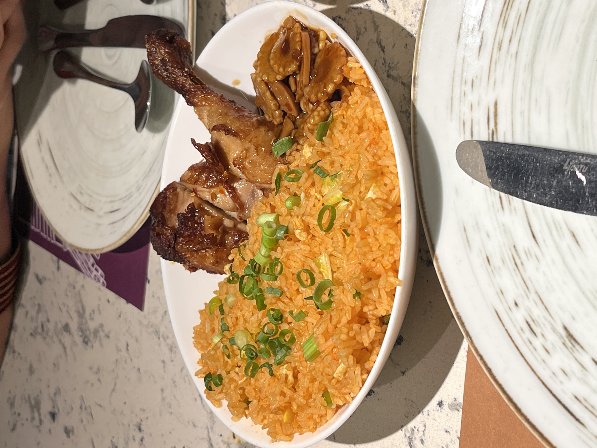 The image shows a plate of food on a table. The food appears to be fried rice with chicken and some mushrooms in a sauce. There are two other plates visible in the background, along with silverware. The table has a speckled, light-colored surface.
Without further information, it is not possible to definitively identify the location as Dhaka, Bangladesh.