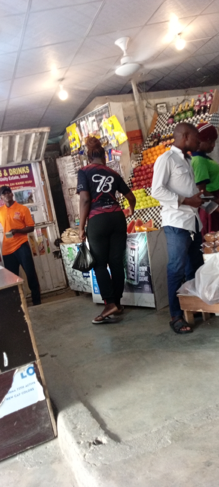 This image captures a scene at an open-air market stall in Aviation Quarters, Nigeria. The stall appears to be selling a variety of items, including drinks and fruit. In the foreground, a woman in black pants and a black t-shirt with a white "B" logo walks away from the camera, carrying a black plastic bag. To her left, a man in an orange shirt and black pants is visible, holding a small item in his hand. Further in the background, behind the woman, a man in a white shirt and blue jeans stands looking to the side, and another person in a green shirt is partially visible.
The setting is a busy market environment with a corrugated metal roof overhead, illuminated by artificial lights. Display shelves are stocked with bottles of drinks and a colorful array of fruits like apples and lemons. A large poster or advertisement with the word "HYPE" is prominently displayed on a stand. There are signs visible with text such as "S & DRINKS," "Unity Estate, Jaba," "NA ZAI KARBI KUDI," "KASHIYA," and "OWN THE GAME." The concrete floor suggests an indoor or covered outdoor space. The overall impression is of a typical market day, with people engaged in shopping or working at the stalls. The lighting suggests it could be daytime, but the indoor setting makes it difficult to determine the exact time of day or weather conditions.
