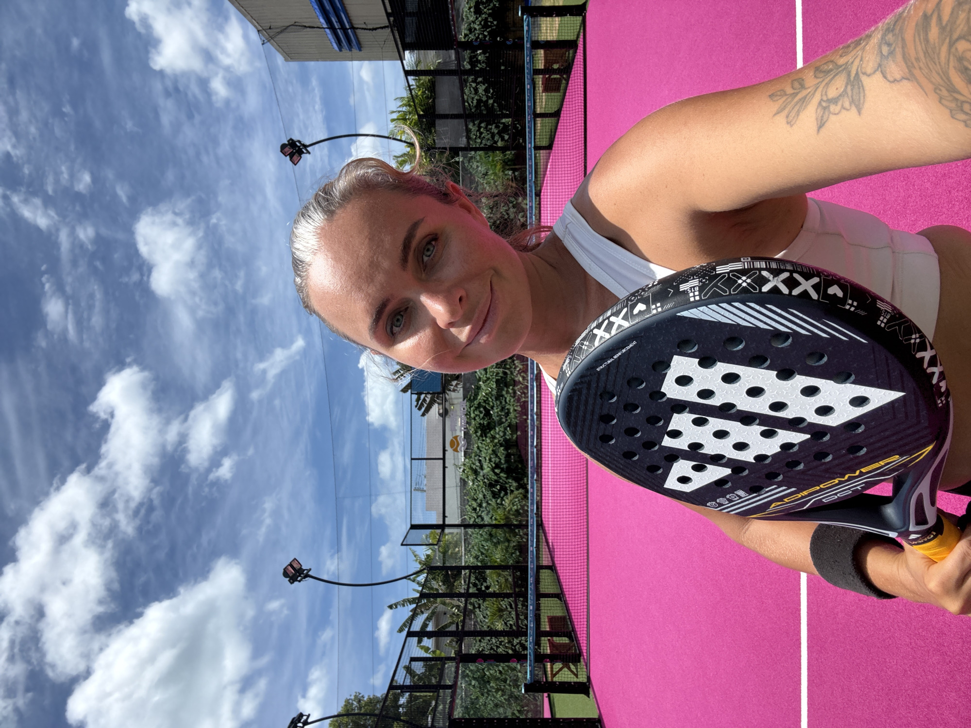 A young Caucasian woman with blonde hair pulled back and a tattoo on her right arm is taking a selfie on a bright pink padel court. She is holding a black and white Adidas padel racket in front of her, with the racket head prominently displayed. She is wearing a white sports bra and a black wristband on her left wrist. In the background, there is a net, fences, lush green tropical vegetation, and buildings. The sky is blue with white, fluffy clouds, and stadium lights are visible above the court. The setting appears to be an outdoor sports facility in the Ko Samui District of Thailand.
