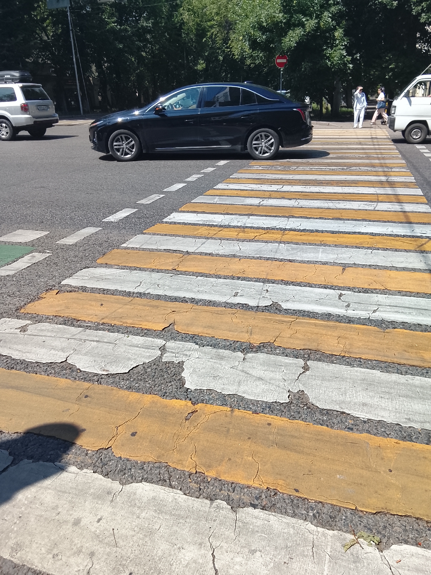 An exterior, eye-level, medium shot captures a pedestrian crossing at an urban intersection. The crossing is marked with alternating yellow and white stripes, showing signs of wear and tear with cracks in the paint. A black sedan is stopped on the road, slightly to the left of the frame, partially obscuring a white SUV in the background. In the distance, on the right side of the pedestrian crossing, two individuals, one in white clothing and the other in darker attire, are walking. A white van is also visible on the right. The scene is set against a backdrop of lush green trees. The overall environment suggests a sunny day in Almaty, Kazakhstan.